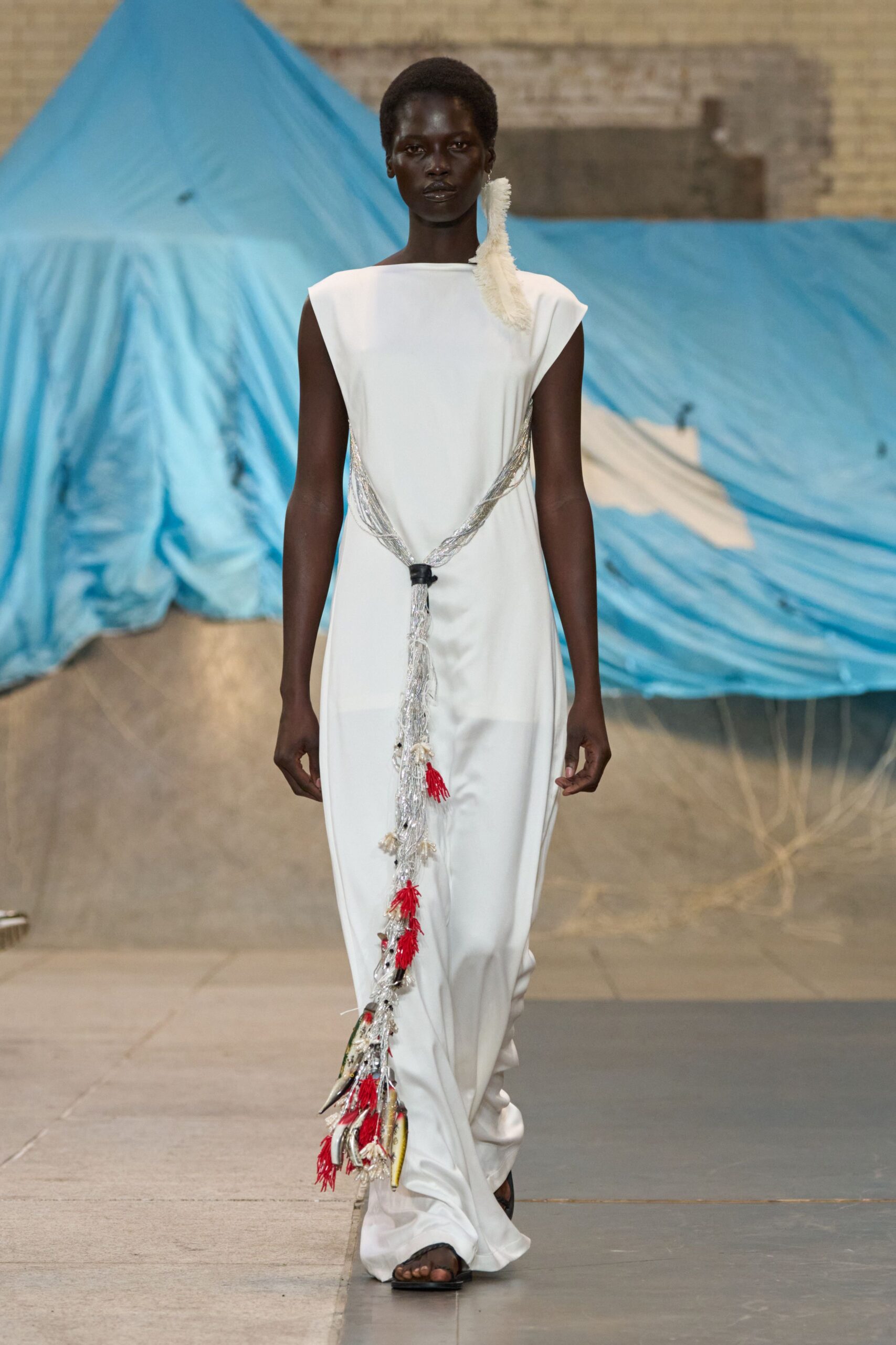 Ivory column dress in fluid crepe, cut with broad cap sleeves. White feather spray rising from one shoulder. Silver cord draped low, knotted with red tassels and shell charms. Flat black thong sandals. Crossing a harbor hall after the blessing boats came in.