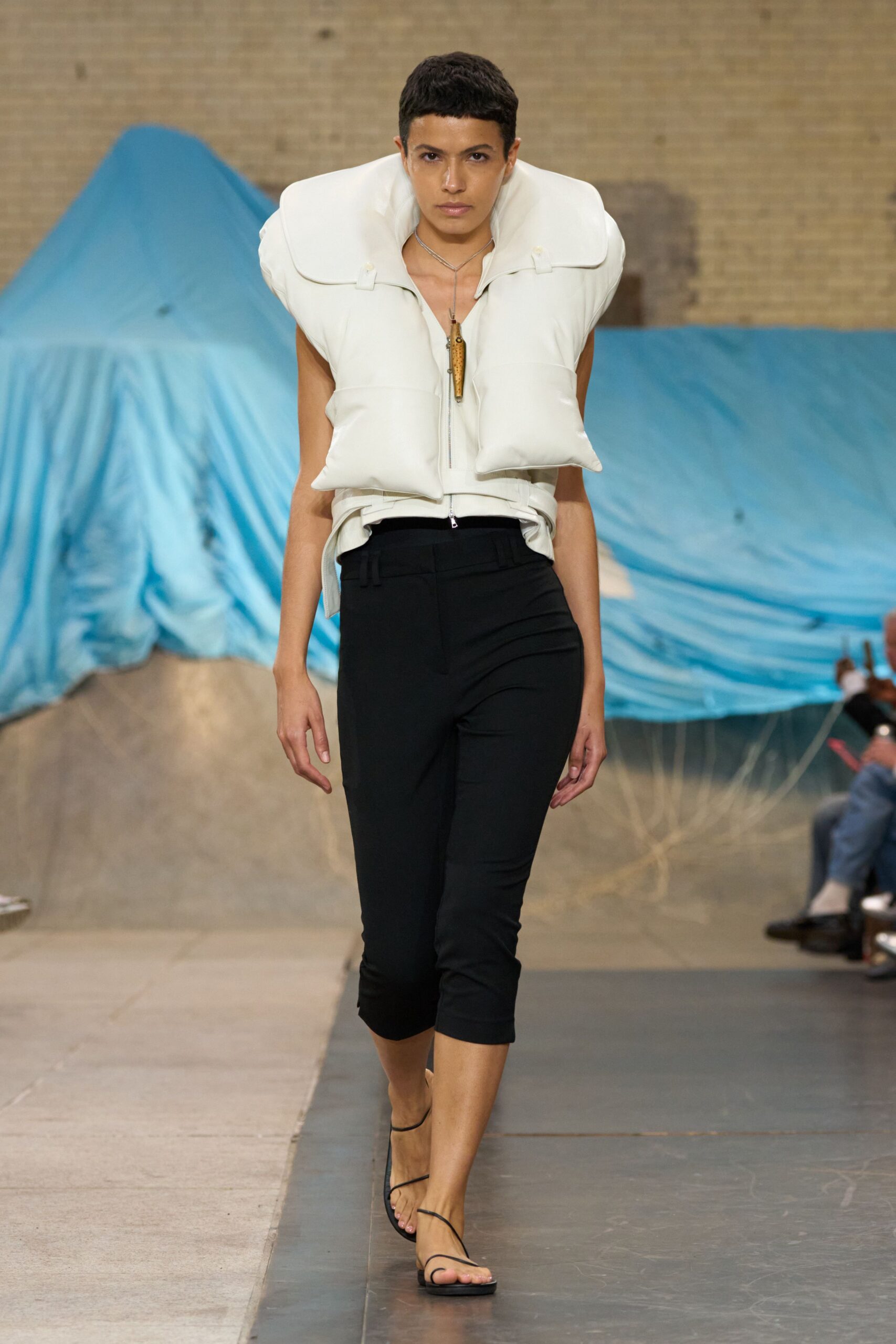 White padded vest with an oversized collar and exposed zip. Long cord necklace hung with a varnished fishing lure. Black slim cropped trousers, pressed creases bending at the knee. Barely-there black thong sandals with wraparound ankle straps. Walking back from the marina with salt drying on your shoulders.