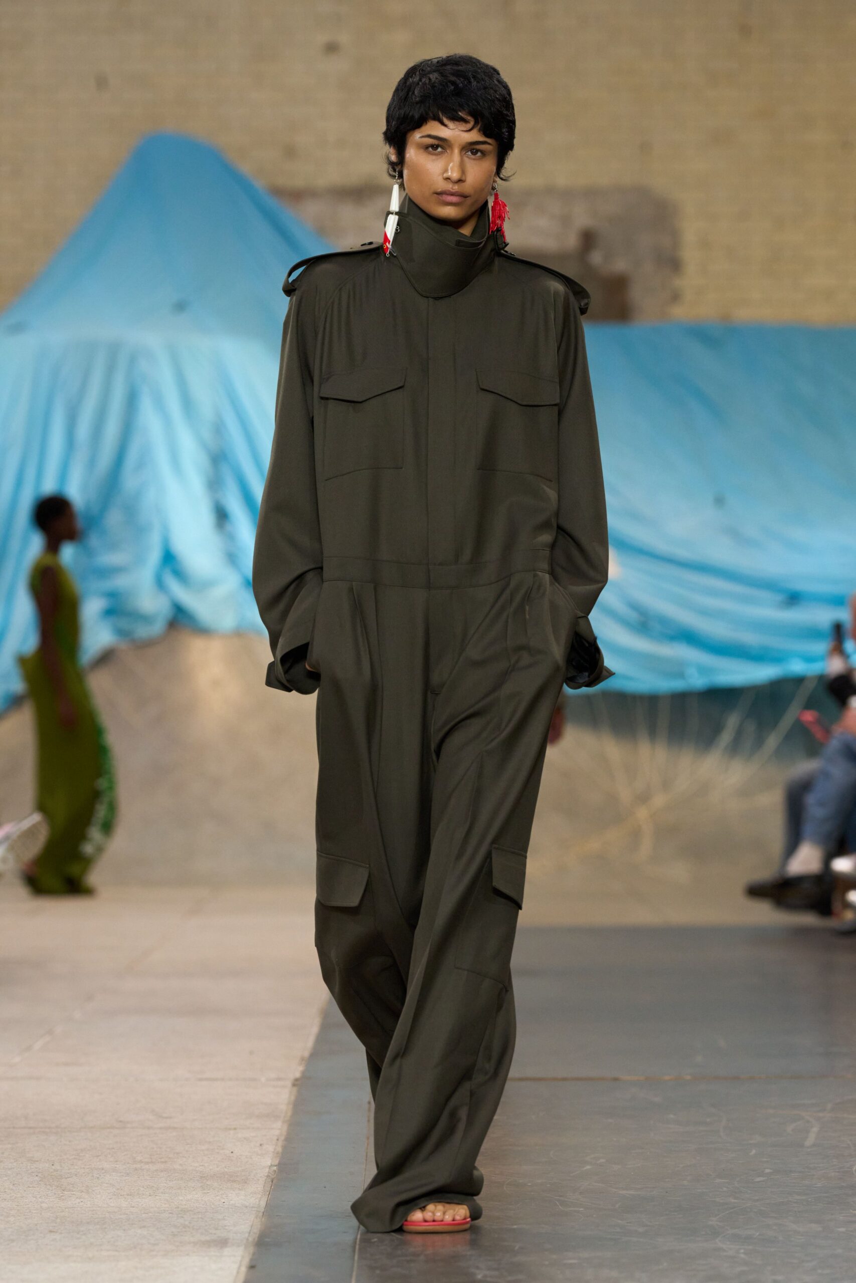 Olive utility jumpsuit in fluid suiting, storm collar rising to the jaw. Mismatched tassel earrings with white spike drops and red thread. Flat thong sandals in hot pink. Walking across an airstrip with tarpaulins snapping in the wind.