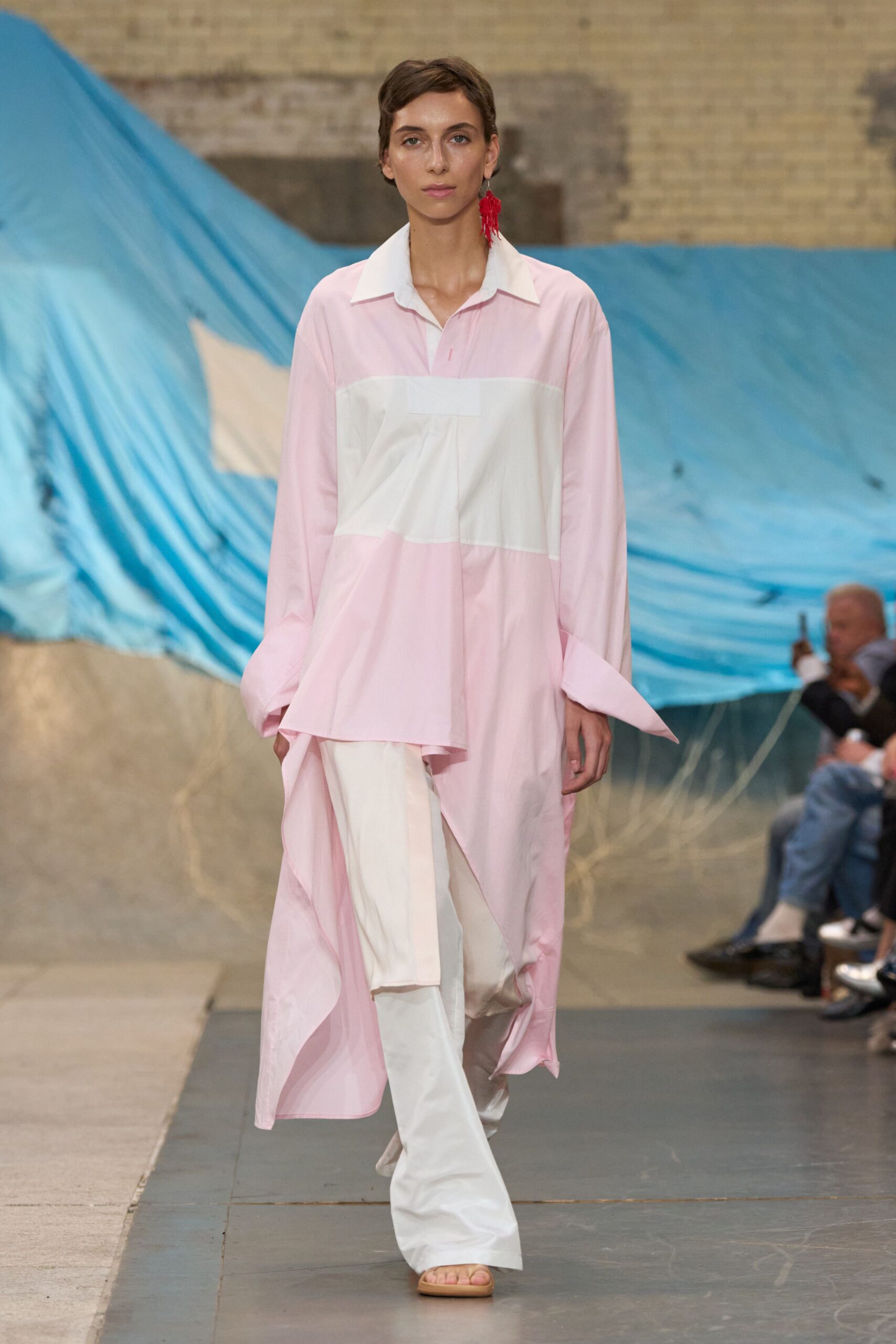 Pale pink cotton tunic shirt with a white bib panel and deep side drape. White wide-leg trousers breaking softly over the sandal. Single red tassel earring at one ear. Flat tan slide sandals. Walking from the guest room to a shaded terrace with wet hair.
