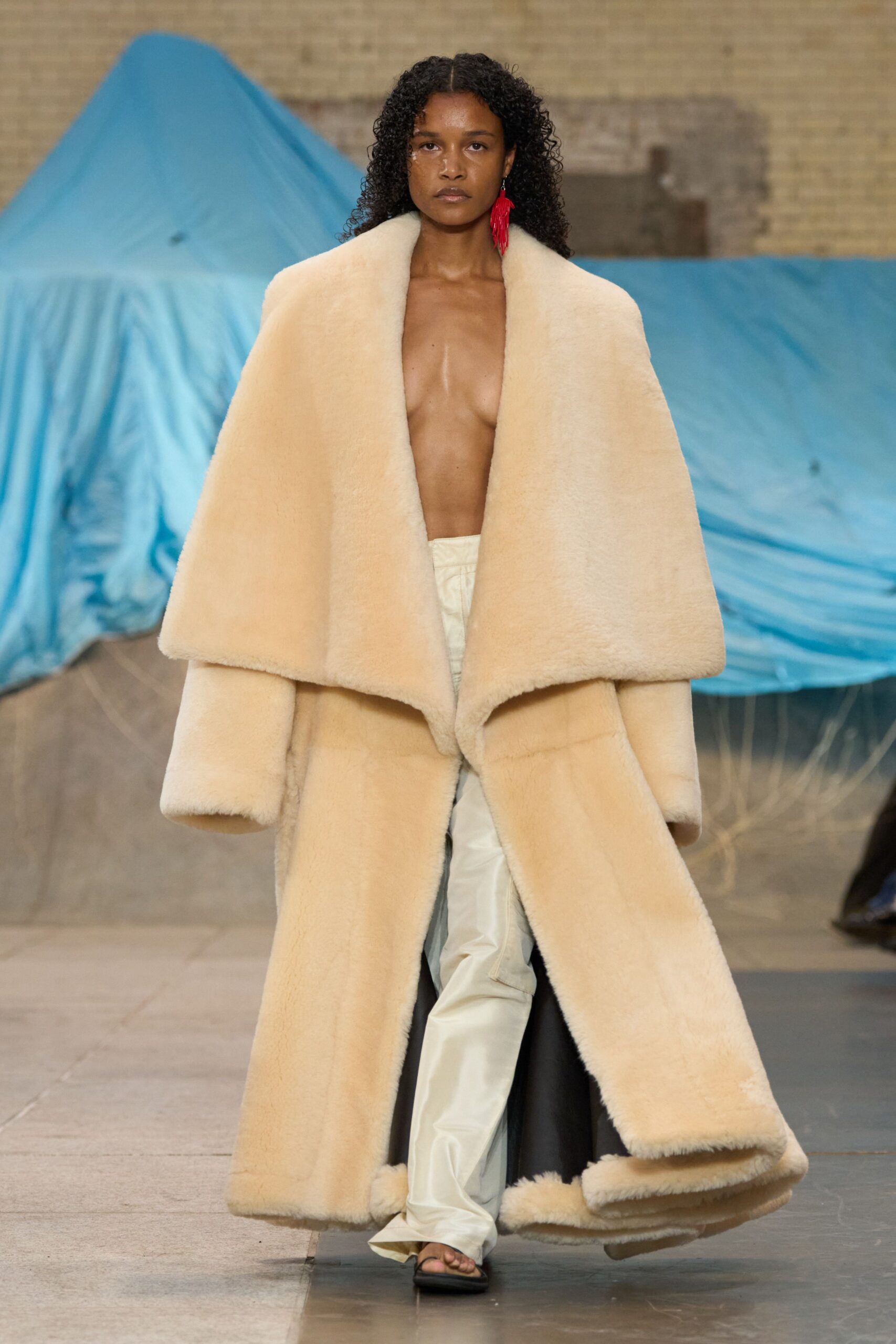 Cream shearling coat with an enormous shawl collar swallowing the shoulders. Single red tassel earring brushing the jaw. Ivory satin trousers, creased and puddling over the sandal. Black thong sandals, barely there. For stepping out of a beach house at dawn wrapped in somebody else's winter.