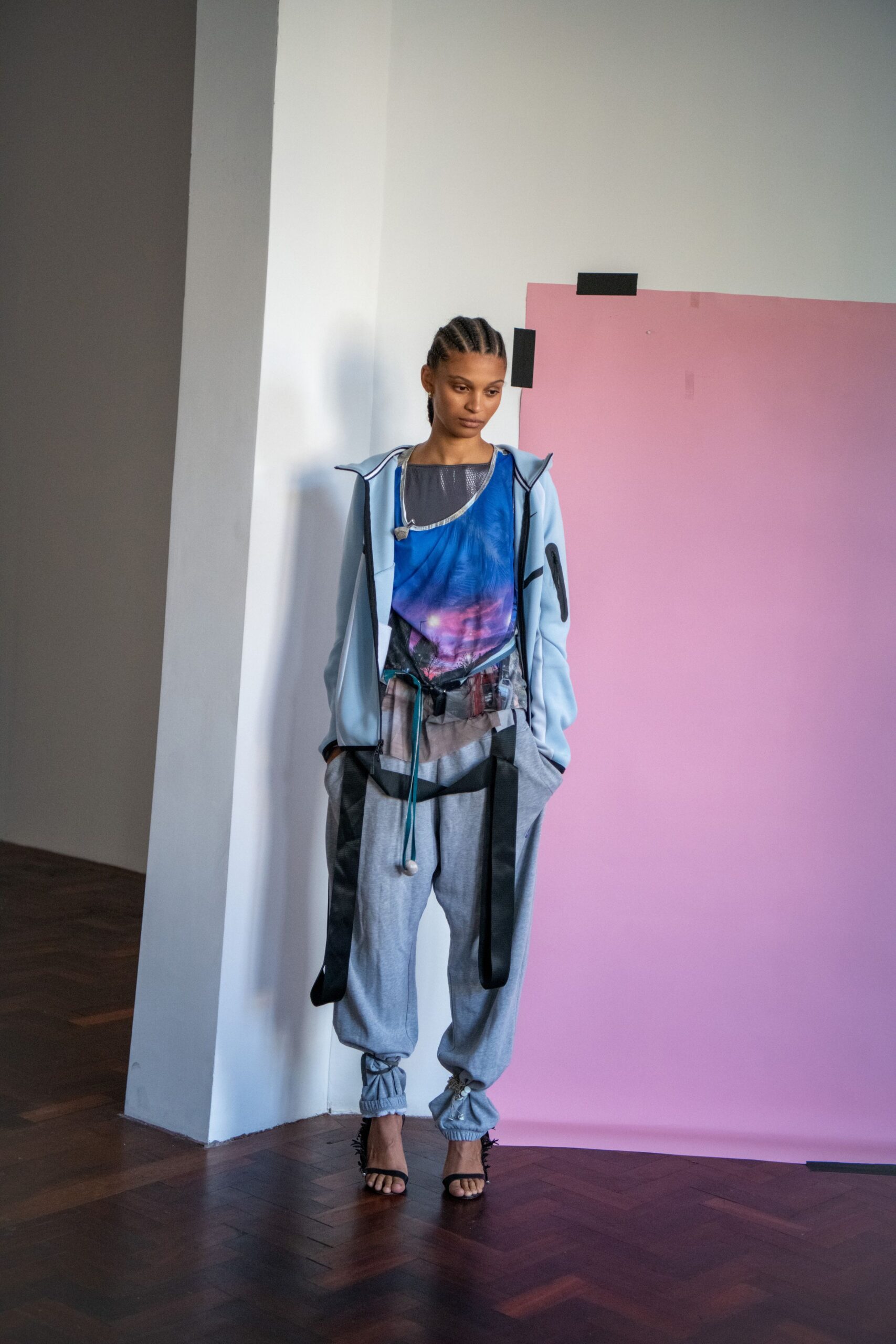 Pale blue zip hoodie traced in black binding, collar standing open. Asymmetric mesh top printed with a violet dusk sky. Grey jersey joggers slung with long black straps and a turquoise drawcord. Black feather-fringed stiletto sandals. Waiting for the printout to finish in an architecture studio at sunset.