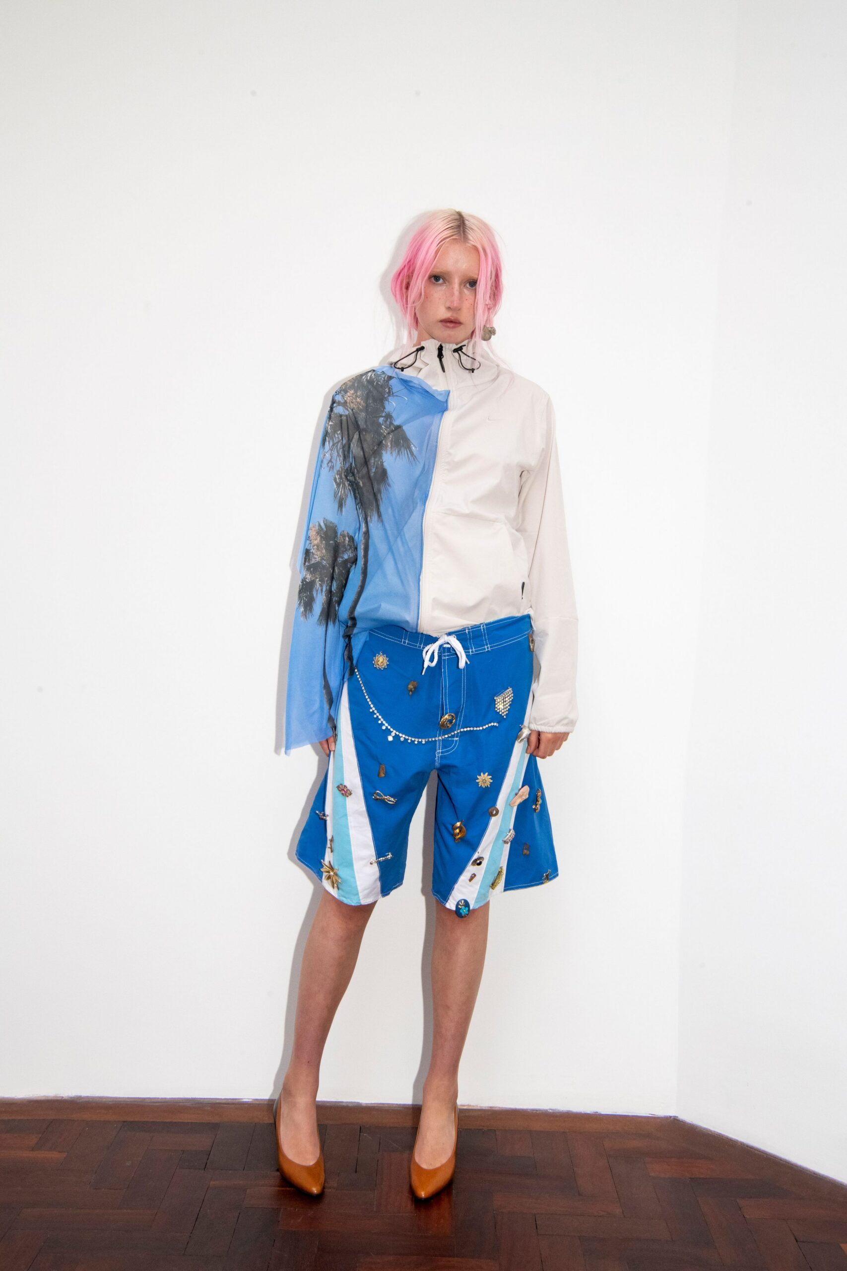 White zip windbreaker with black bungee toggles and a blue palm-print panel. Electric-blue board shorts, white topstitching and souvenir charms pinned everywhere. Tan leather pumps with a sharp almond toe. Hotel balcony after swimming, still wearing the gift-shop trinkets.