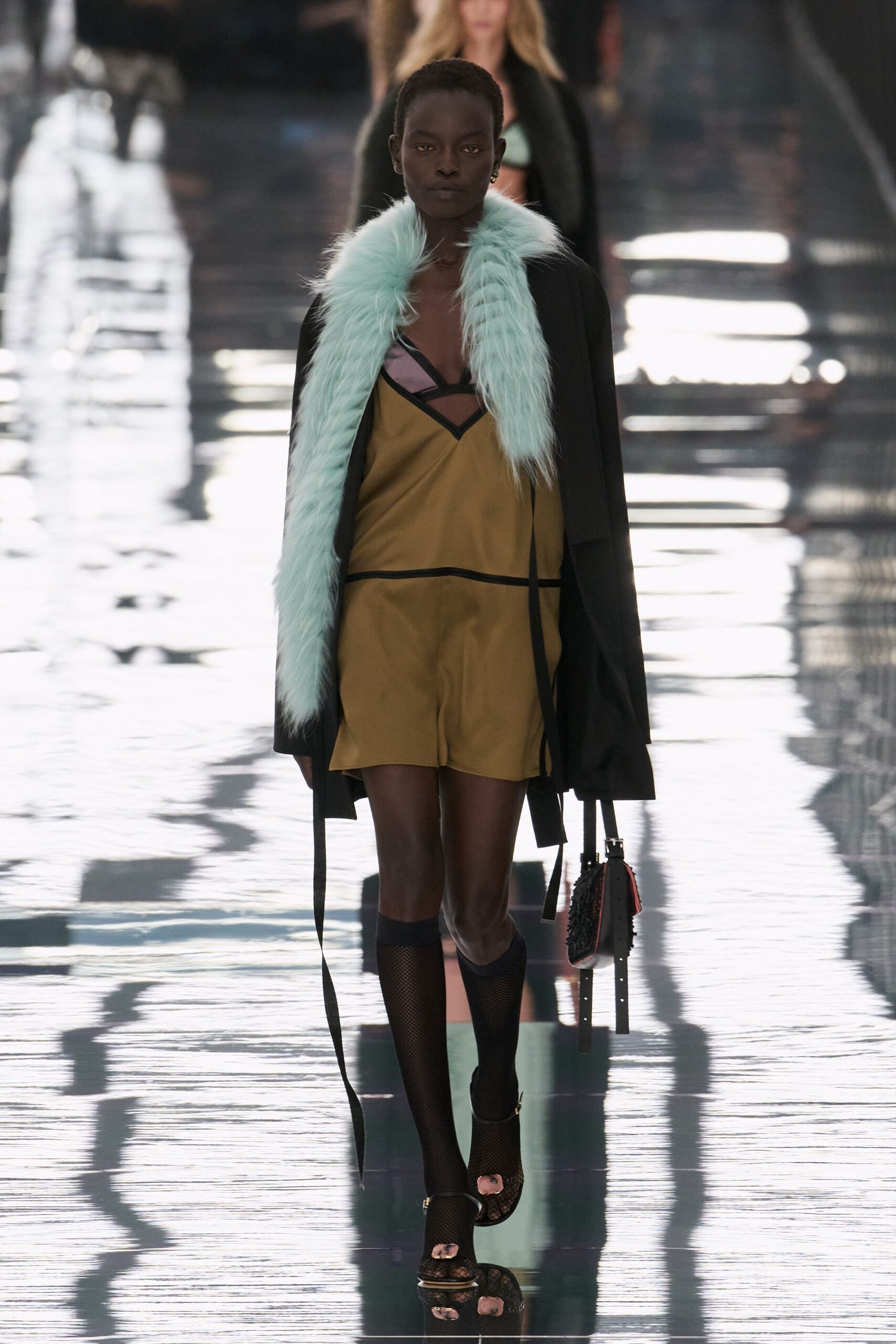 Black wrap coat edged in pale mint long-pile fur. Ochre slip mini with black binding and lilac bra inset. Small black shoulder bag with red sides and dangling straps. Black fishnet knee socks. Black ankle-strap sandals with a pink padded toe piece. Crossing a hotel lobby after rain, keycard already in hand.