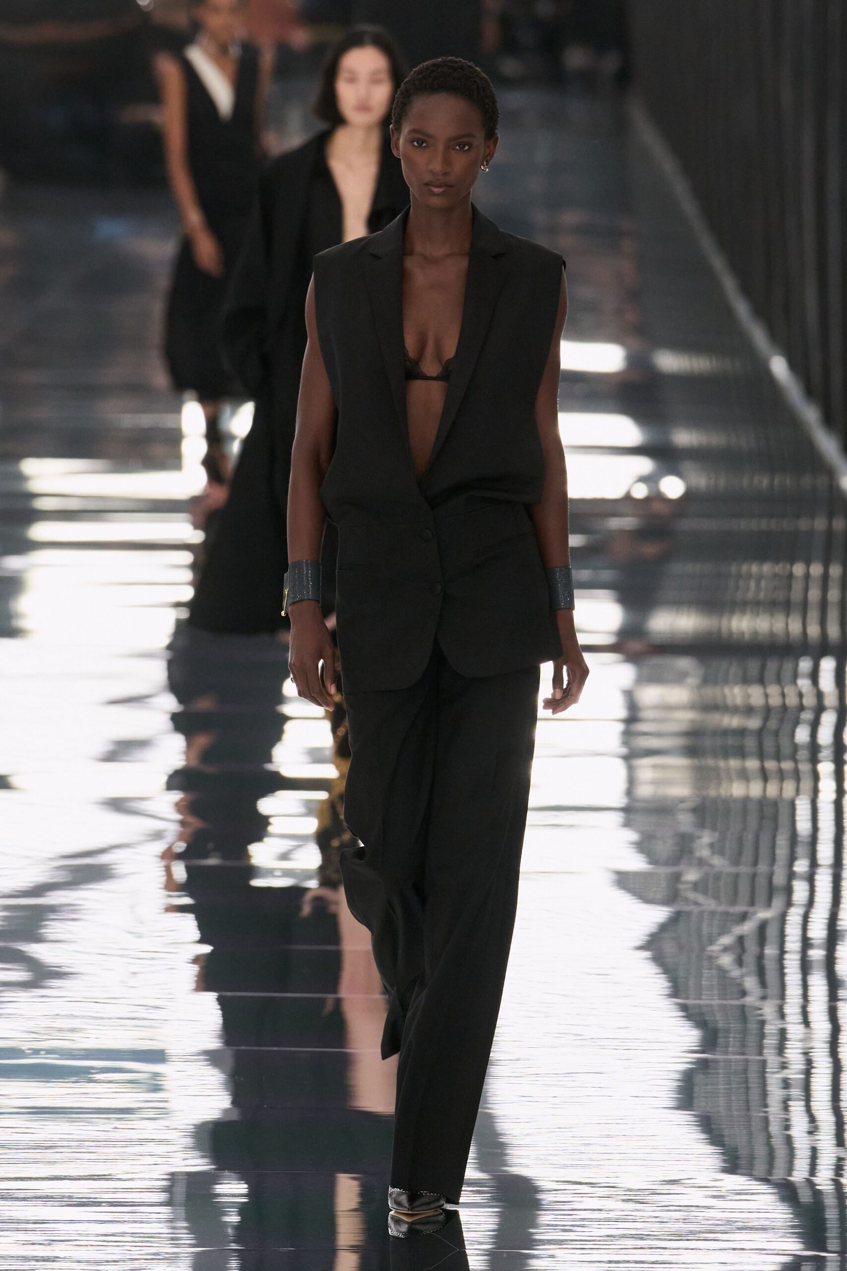 Black lace bralette peeking through a plunging opening. Long black blazer with padded shoulders and two low buttons. Loose black trousers breaking softly over the shoe. Wide gunmetal cuffs on both wrists. Pointed black heels with a pale metal cap. Walking the hotel corridor after midnight, carpet swapped for mirrored stone.