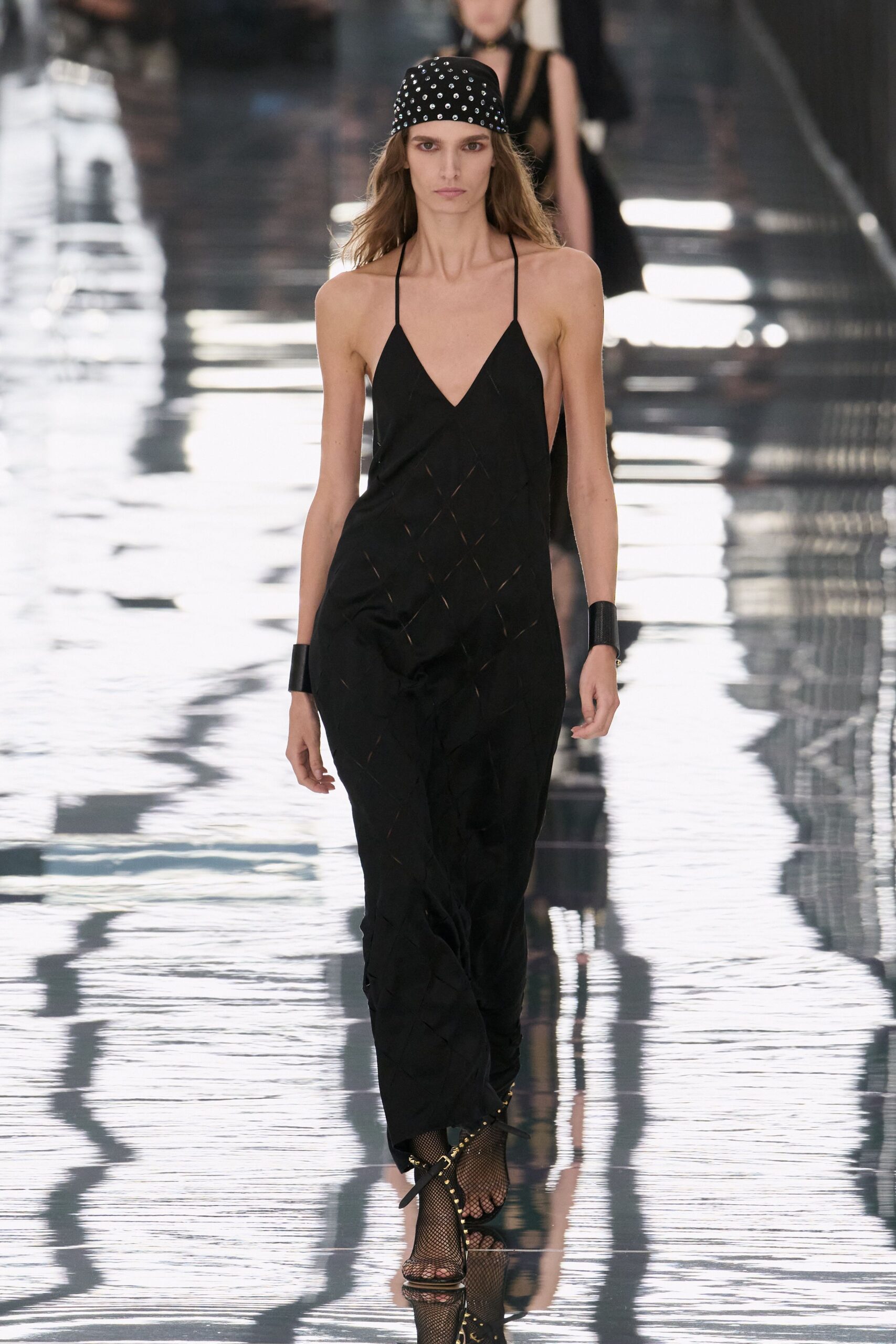 Black headscarf studded all over with tiny silver domes. Black halter jumpsuit in liquid crepe, cut through with narrow diamond slits. Wide black leather wrist cuffs. Fishnet ankle boots laced up the front with gold eyelets. Walking a wet concourse after midnight, lights breaking under every step.