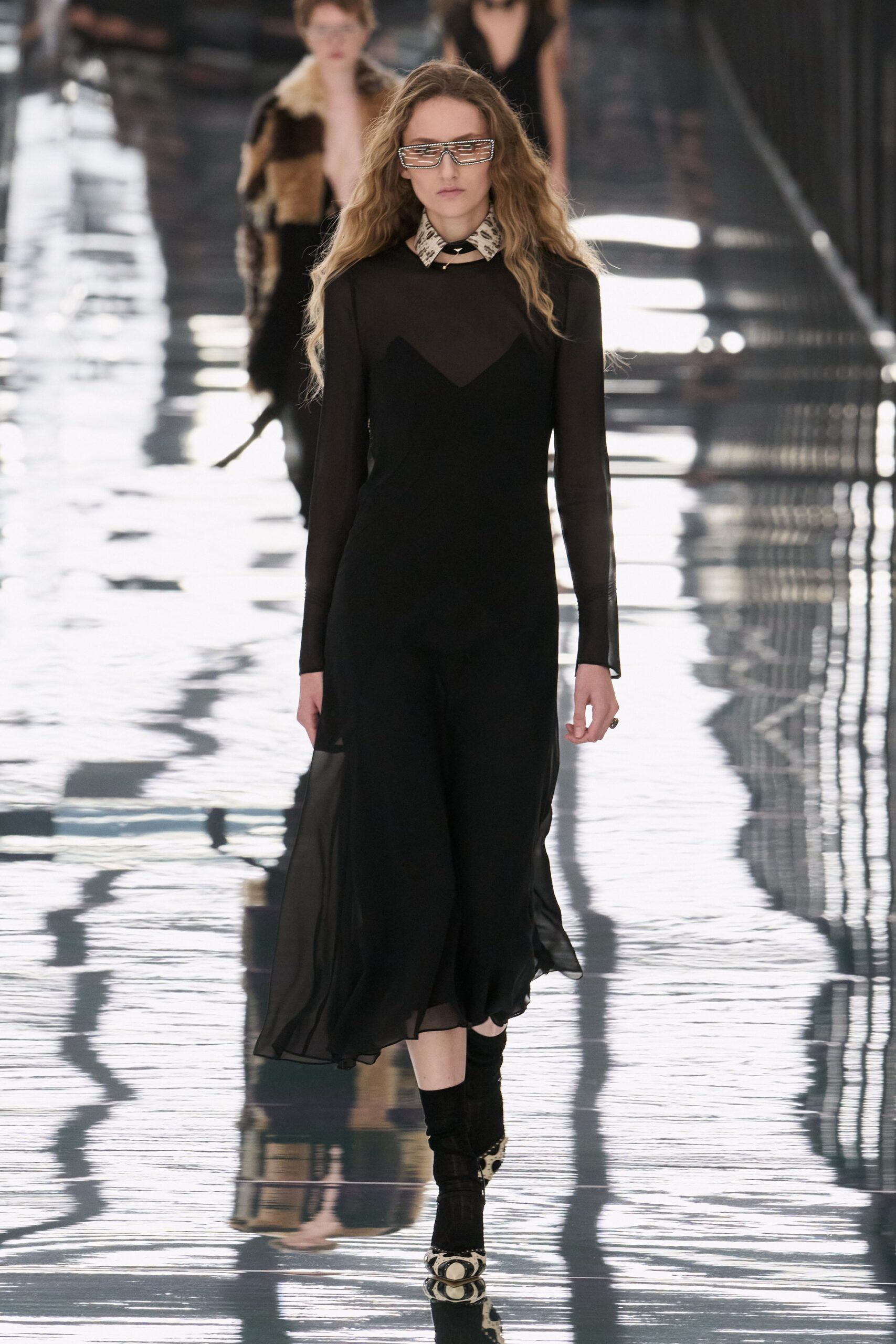 Rhinestone slat visor sunglasses masking the eyes. Cream python-print scarf knotted close with a tiny spike choker. Black sheer mesh dress over a sweetheart slip, hem floating unevenly. Slouchy black ankle boots. Python cap-toe pumps peeking out beneath. Crossing a hotel atrium after rain, marble floor still holding the light.