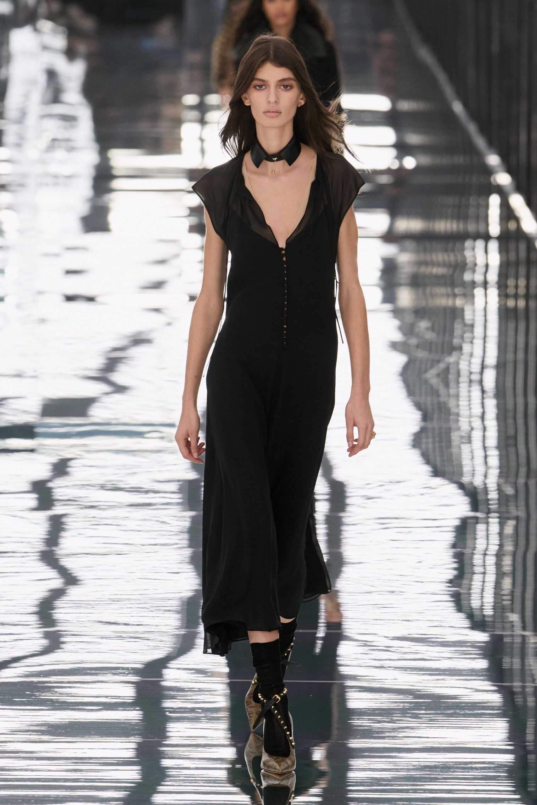 Black leather collar with a tiny gold stud at the throat. Sheer black georgette gown, plunging front laddered with narrow connectors. Black ankle socks. Pointed heels with laced gold eyelets climbing the vamp. Crossing a glassy footbridge after rain, river light flickering underfoot.