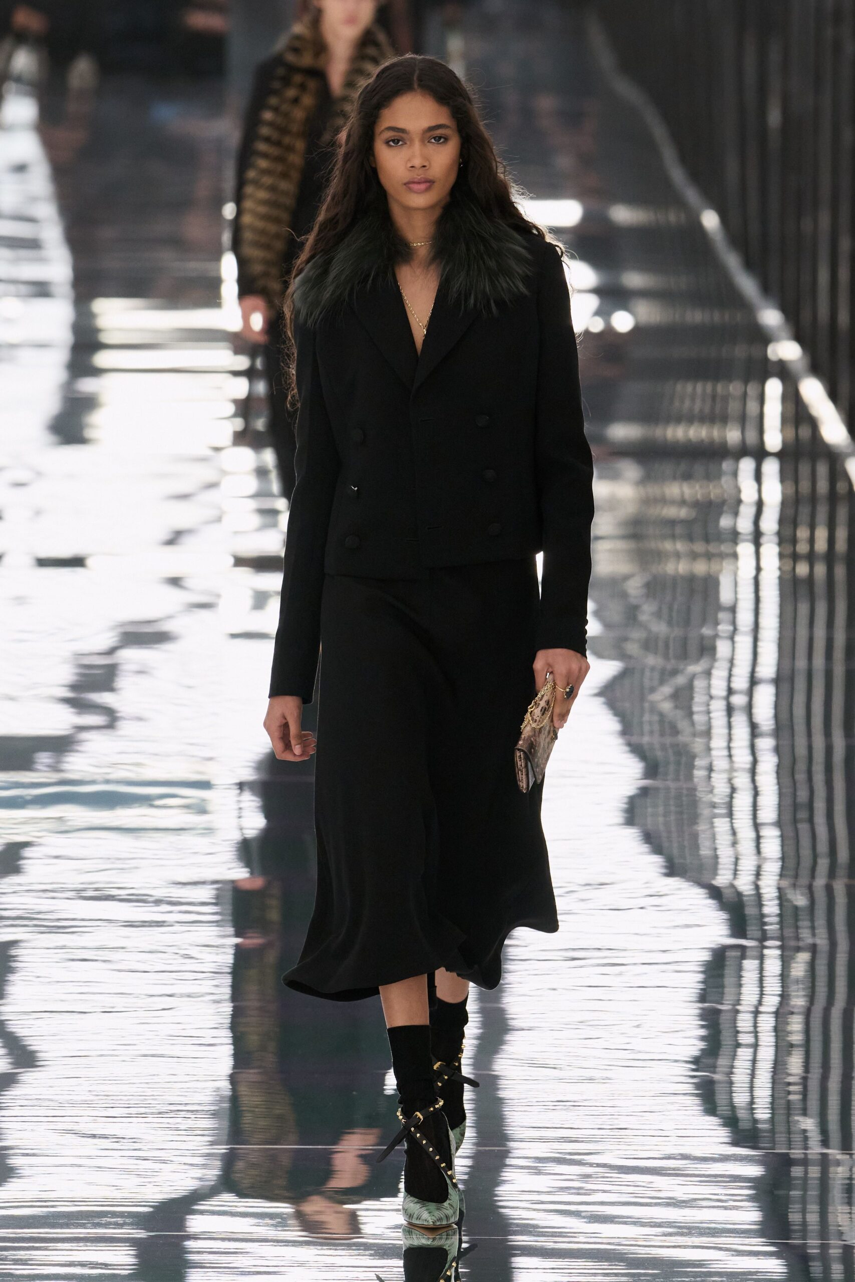 Black double-breasted blazer with a dense inky fur collar. Bias-cut black midi skirt, hem swinging just off the shin. Small python clutch with a gold frame clasp. Pale green pointed pumps strapped with black ties and studs. Black ankle socks. Crossing a hotel atrium before breakfast service starts.