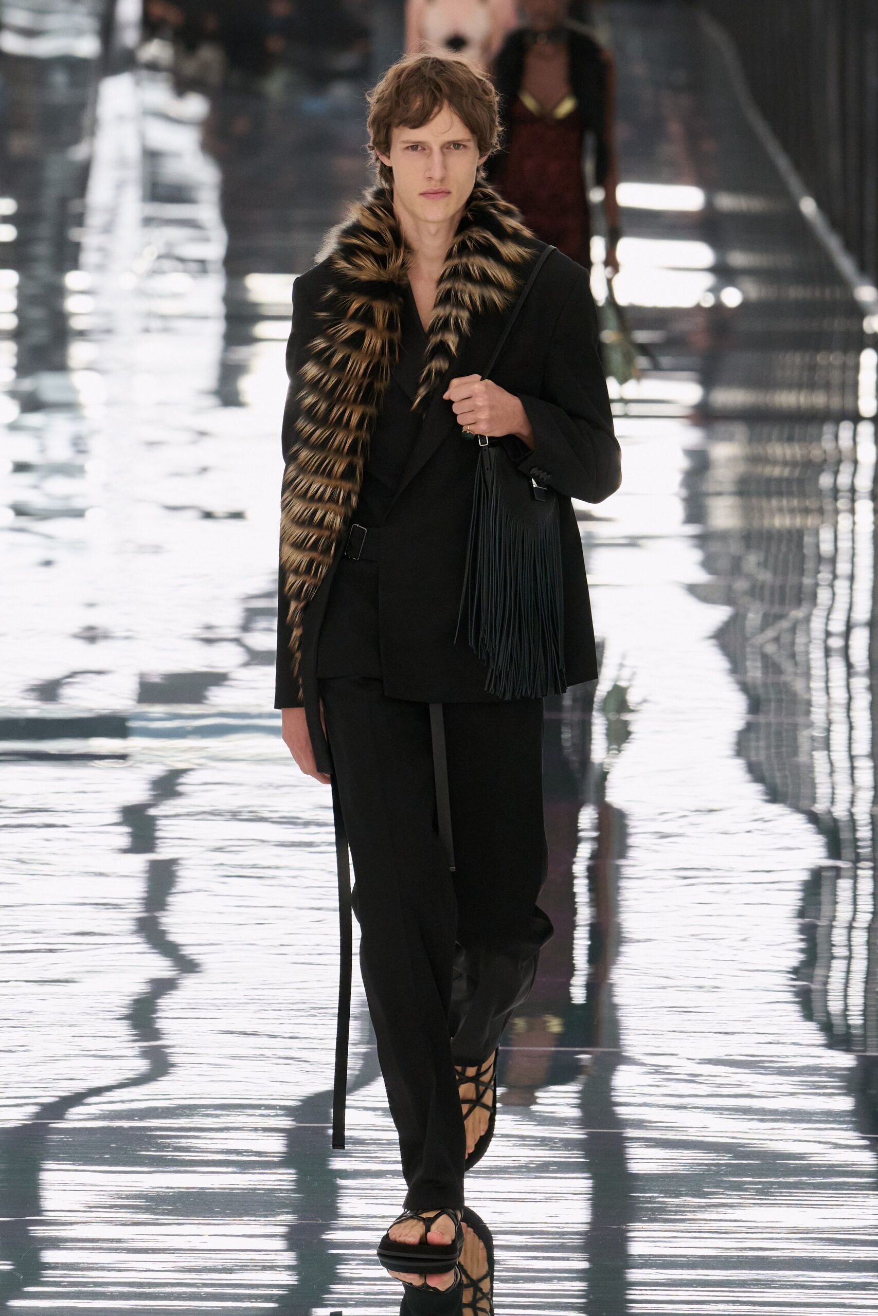 Long striped fur scarf, one tail dropping almost to the shin. Black wrap blazer cinched with a side buckle and loose strap. Straight black trousers breaking softly over the foot. Black shoulder bag with dense leather fringe. Flat black sandals laced across the toes. Crossing a hotel atrium just after the rain stopped outside.