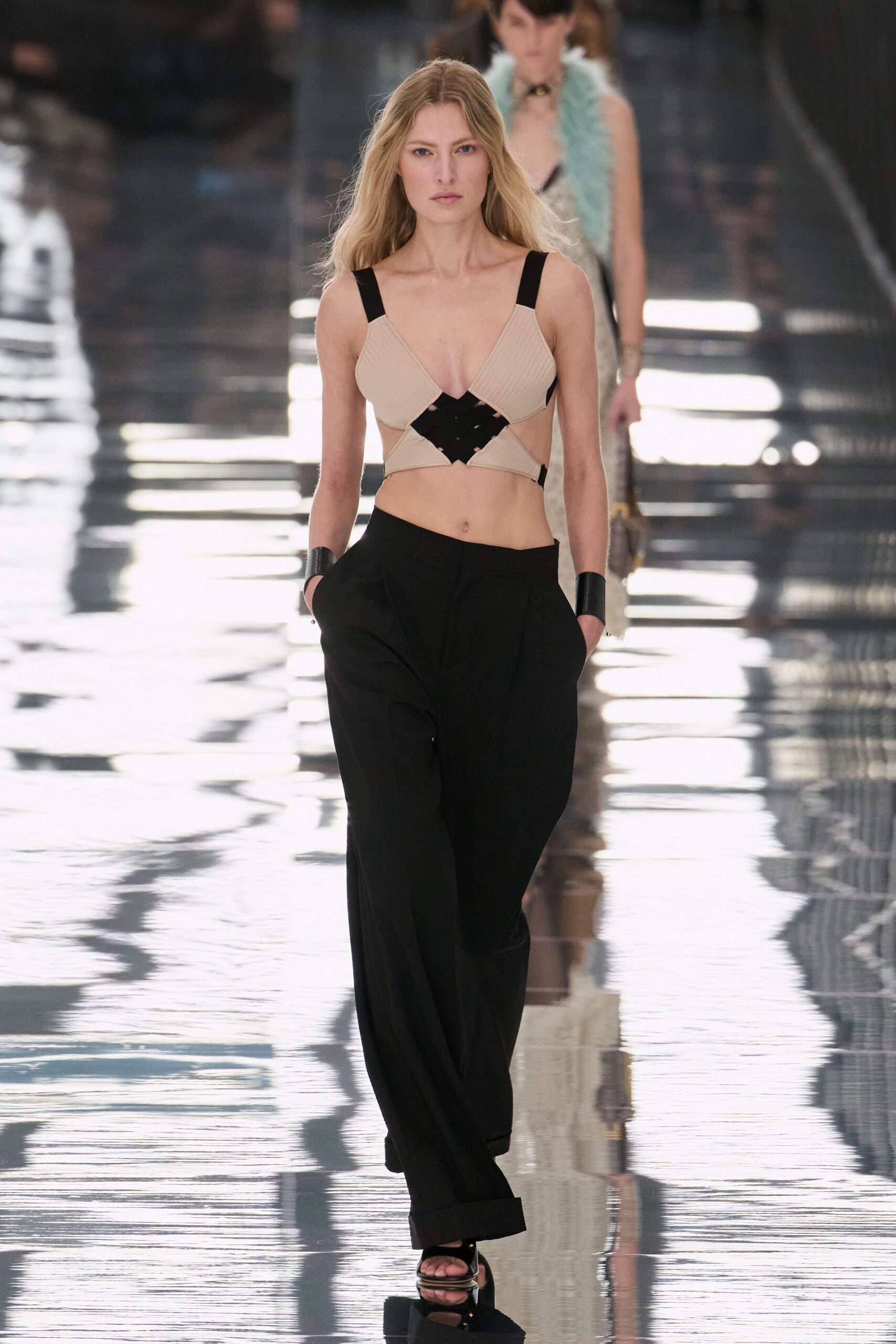 Sand ribbed knit top, black lattice inset bridging the bare midriff. Black fluid wide-leg trousers with deep pleats and turned cuffs. Black leather wrist cuffs, rigid and glossy. Black heeled sandals with a square open toe. Crossing a hotel lobby while the floor is still wet from mopping.