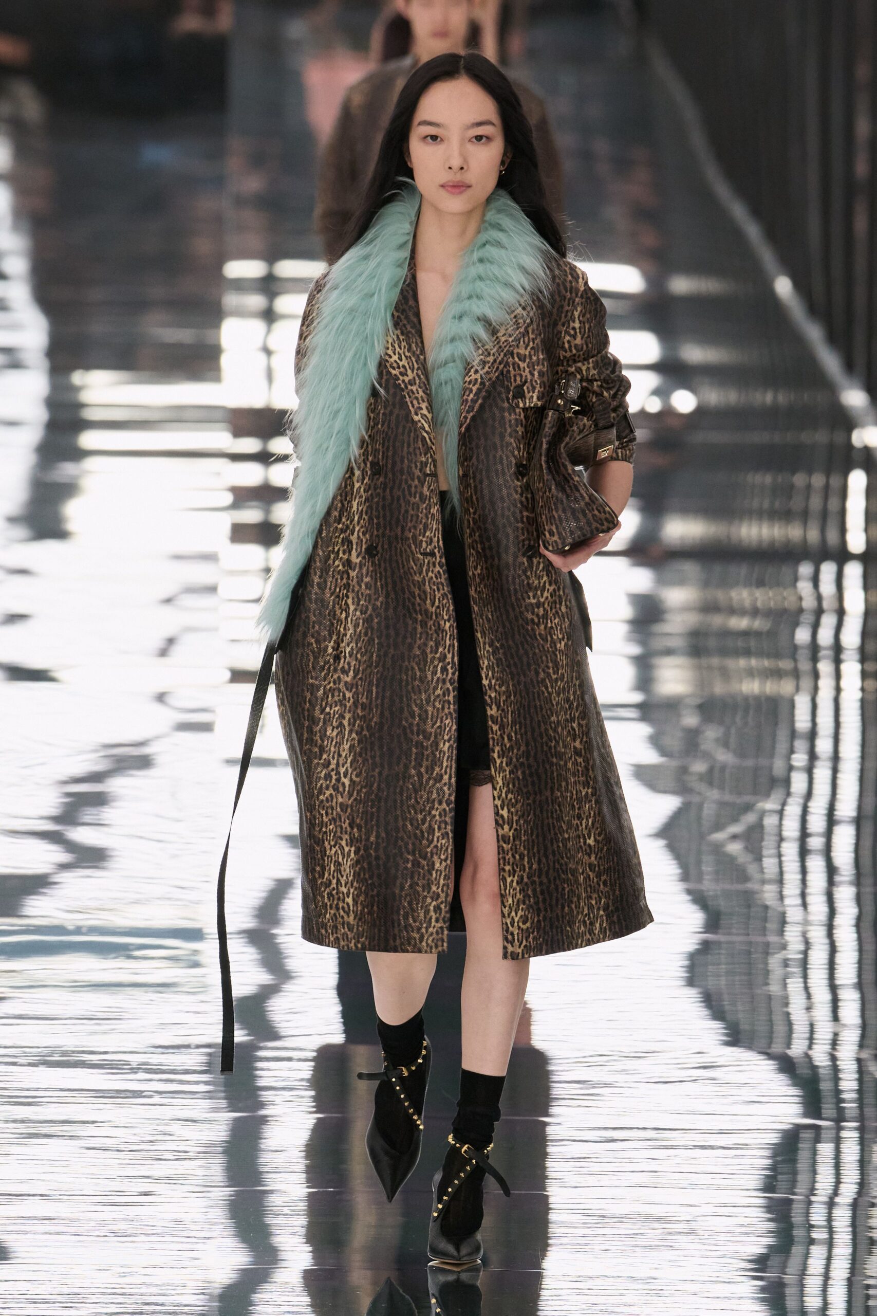 Long leopard coat with a pale aqua fur lapel and loose belt tail. Black slip dress opening to a narrow lace-edged slit. Leopard shoulder bag tucked under the arm with a gold clasp. Black ankle socks. Pointed black heels strapped in gold-studded bands. Crossing a hotel atrium after rain, carrying only what fit under your arm.