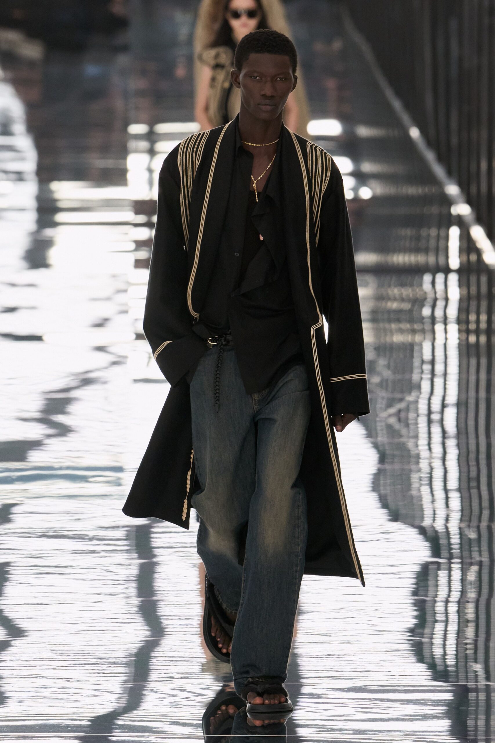 Long black coat traced with narrow ivory cord at every edge. Black blouse with a loose ruffle dropping through the open placket. Faded indigo wide-leg jeans, pooled and soft through the break. Two fine gold chains, one hanging in a slim drop. Black slide sandals. Crossing a hotel terrace just after the pool has been cleared.