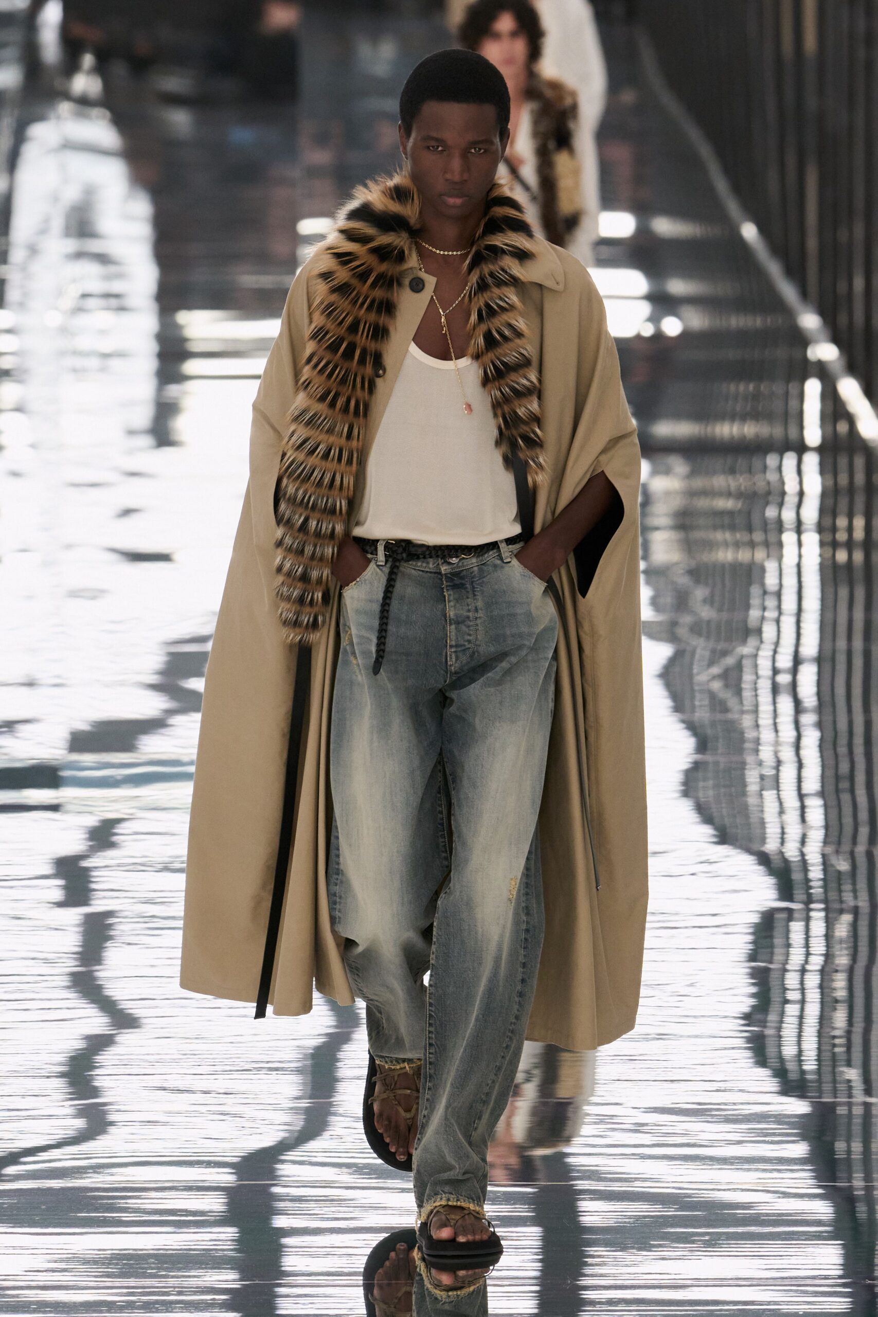Sand cape with a long striped fur panel hanging off one shoulder. Loose ivory tank, cut low and scooped at the chest. Fine gold lariat with a small pink drop. Washed blue jeans, hems shredded into pale fringe. Brown leather sandals on thick black soles. Crossing a marina walkway after noon, water flashing hard off the planks.