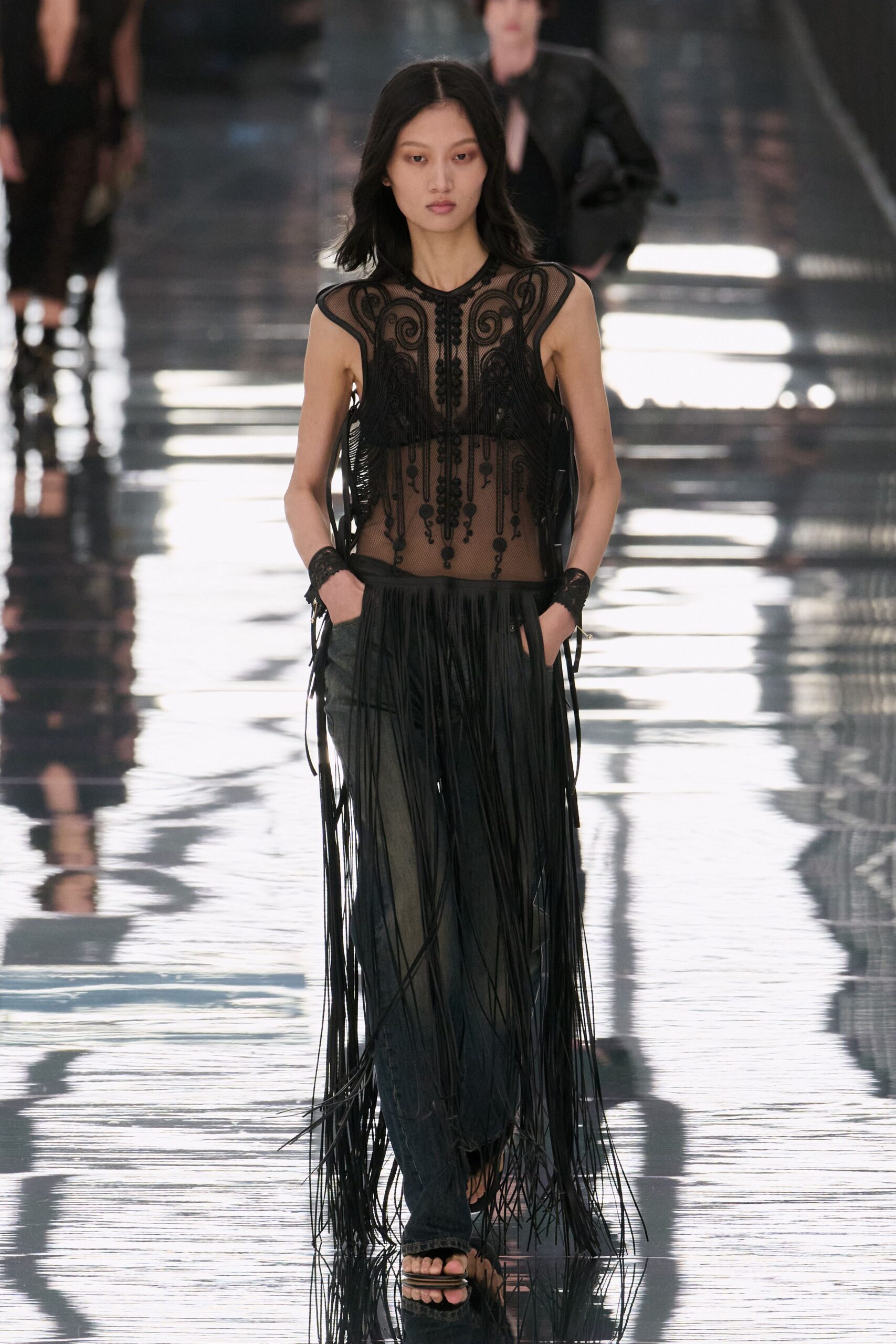 Sheer black mesh top, soutache scrollwork and beaded drops framing the torso. Waist-hung leather fringe falling nearly to the floor. Washed black jeans, loose through the leg. Black lace wrist cuffs. Flat black sandals with stacked sole layers. Walking across a hotel lobby after midnight while the piano is still going.