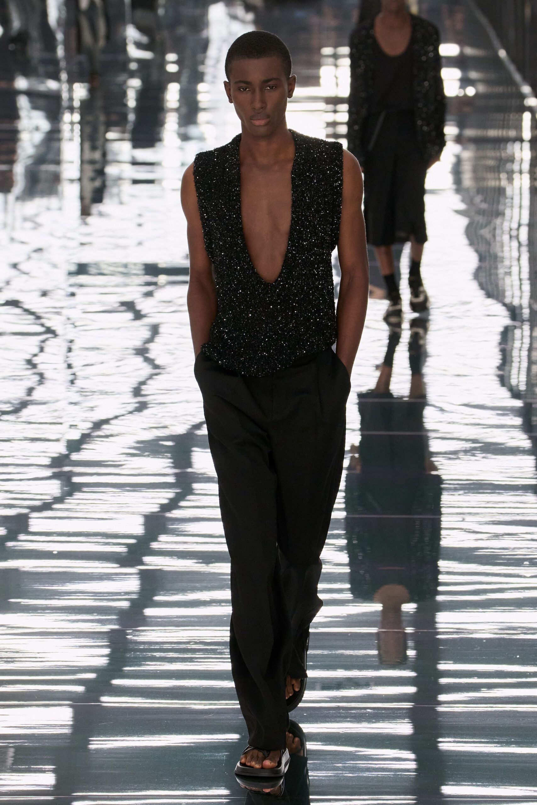 Black beaded sleeveless top with a plunge dropping nearly to the navel. Loose black trousers with soft front pleats and hands sunk deep. Flat black thong sandals, thick sole under bare toes. Crossing a hotel atrium after midnight, floor still wet from mopping.
