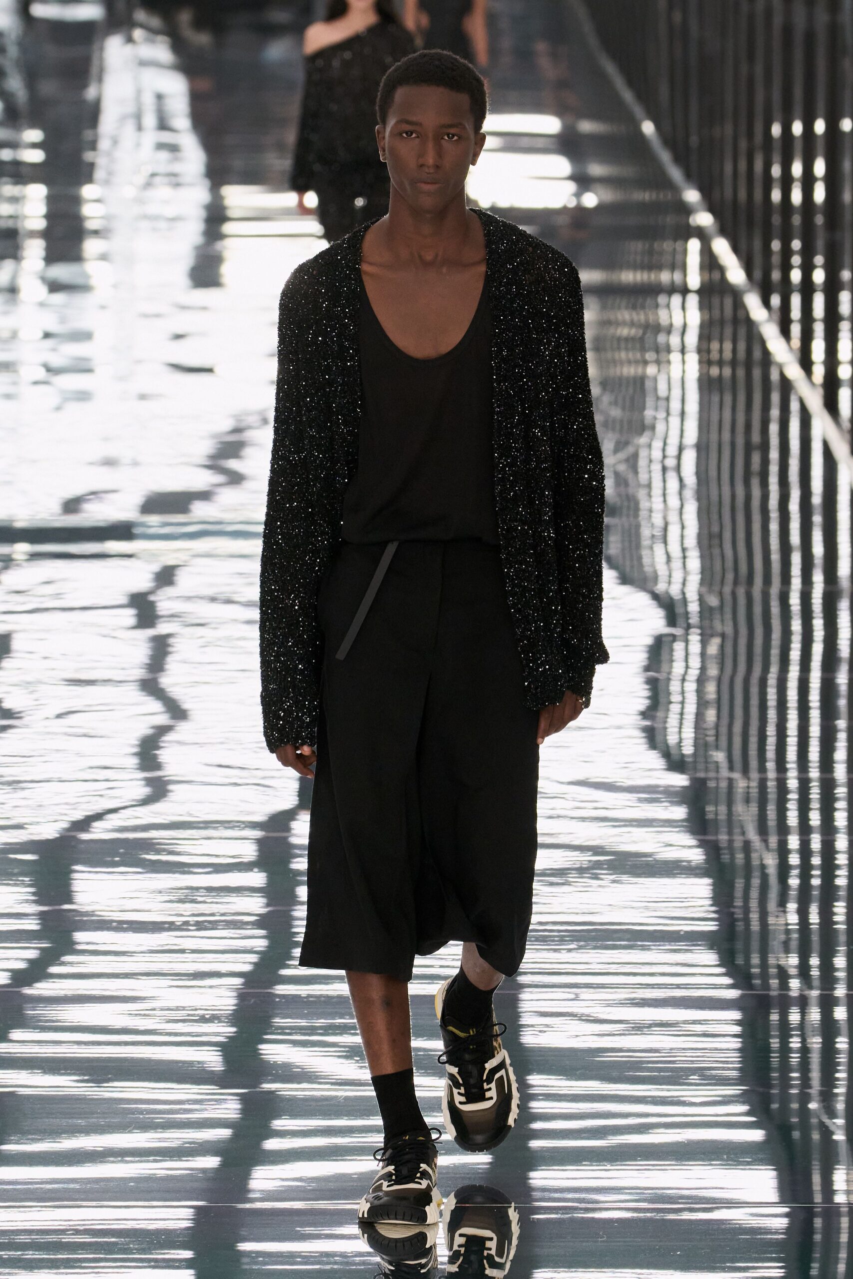 Black cardigan densely crusted with tiny sequins, hanging open and loose. Deep scoop black tank in sheer jersey. Black cropped trousers with a long belt tab dropping from the waist. Black ankle socks. Chunky black sneakers with white rubber tracing the sides. Crossing a wet concourse after midnight cleaners have already started.