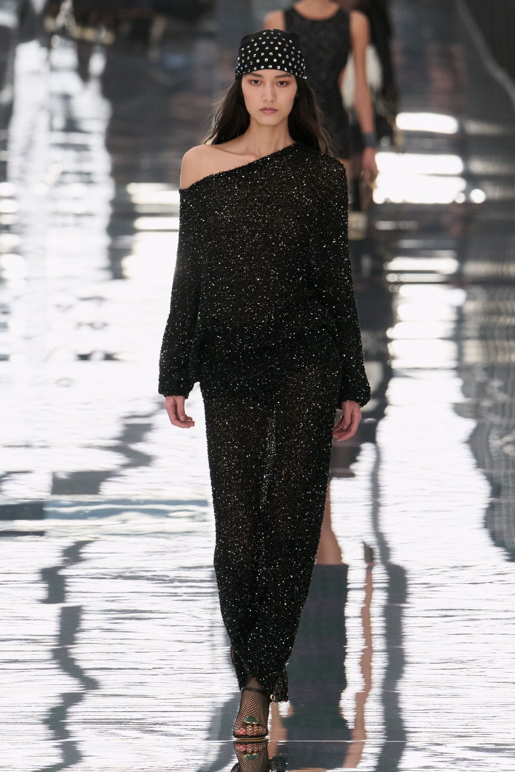 Black satin bandana set with neat rows of silver studs. Black sequined knit dress, cut off one shoulder with balloon sleeves. Black fishnet tights. Peep-toe heels with a green jewel buckle at the vamp. Crossing a hotel atrium after midnight, floor still wet from mopping.