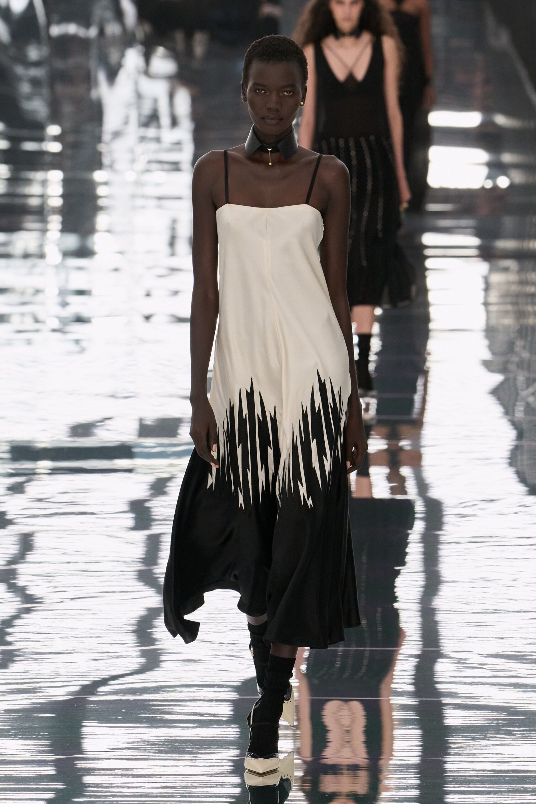 Wide black leather choker with a tiny gold drop at center. Ivory slip dress with razor-cut black shards climbing from the hem. Black ankle boots with pale folded leather caps at the toe. Crossing a hotel lobby just after the marble floors were mopped.