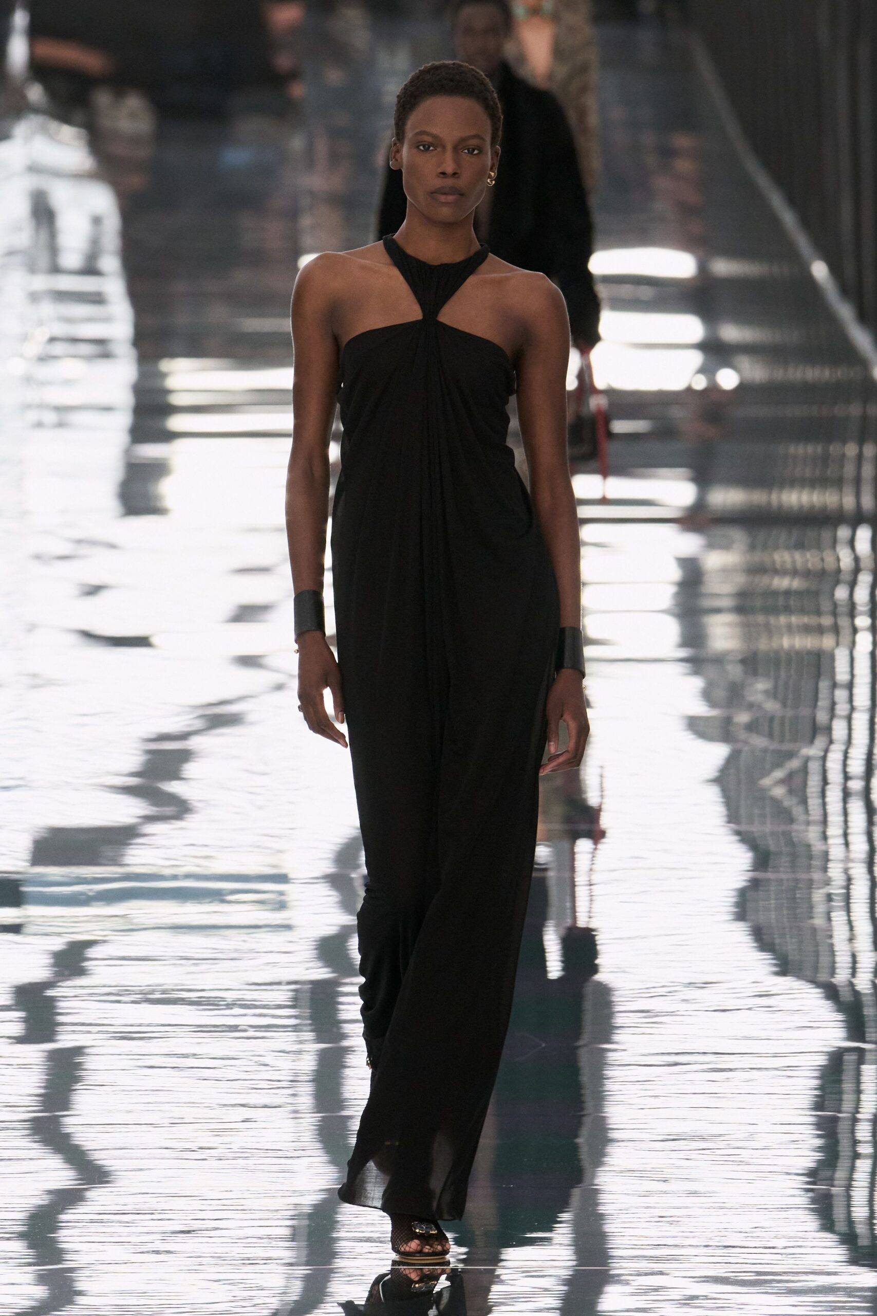 Black jersey column gown, twisted halter knot dropping into a gathered front panel. Wide black leather cuffs on both wrists. Barely there sandals with fishnet over the toes. Crossing a hotel atrium after midnight while the floor still smells mopped.