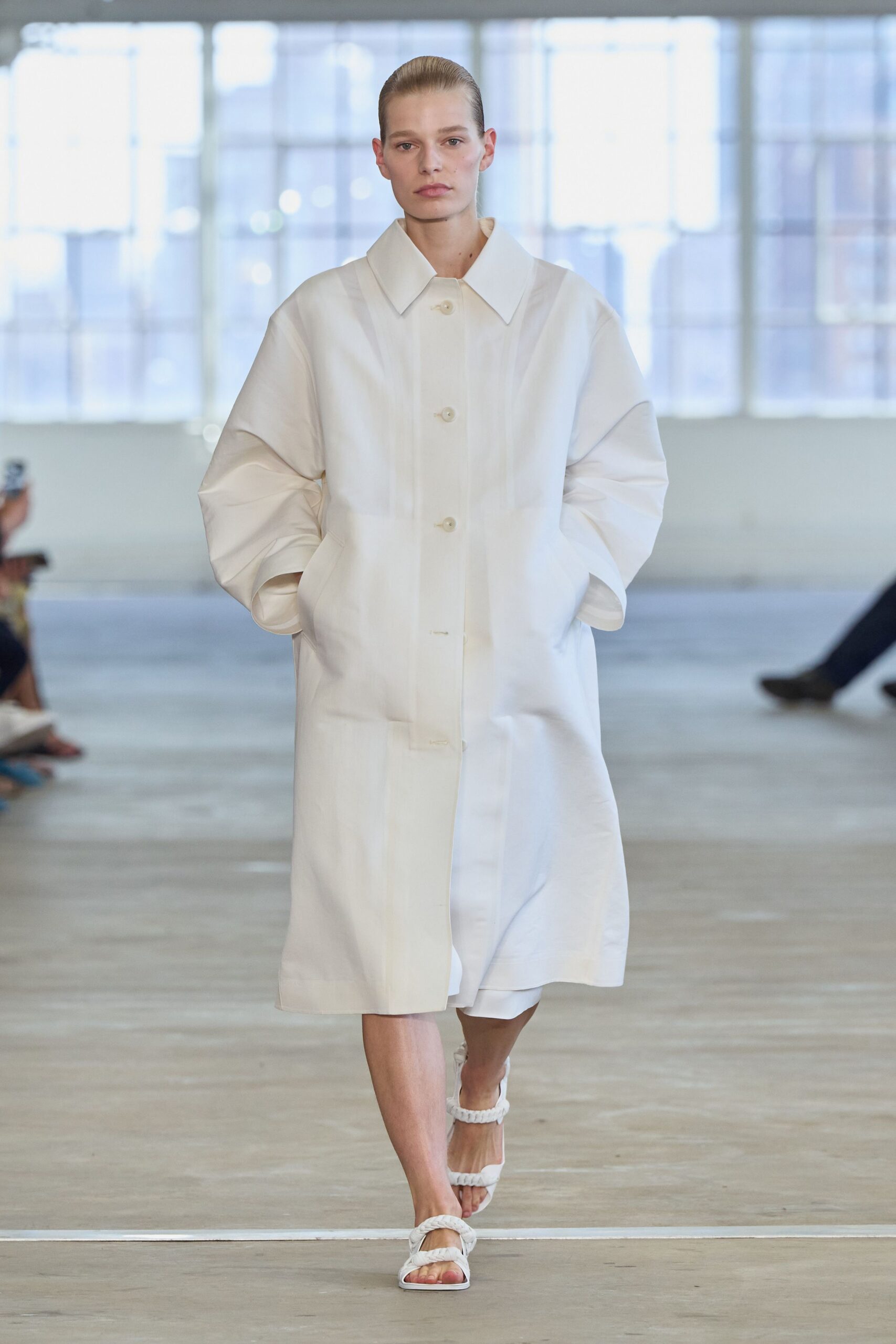 Oversized ivory coat with a broad collar and softly ballooned sleeves. White knee-grazing shorts peeking below the coat hem. White sandals with thick braided straps across toe and ankle. Walking through a near-empty museum before the air-conditioning fully wakes up.
