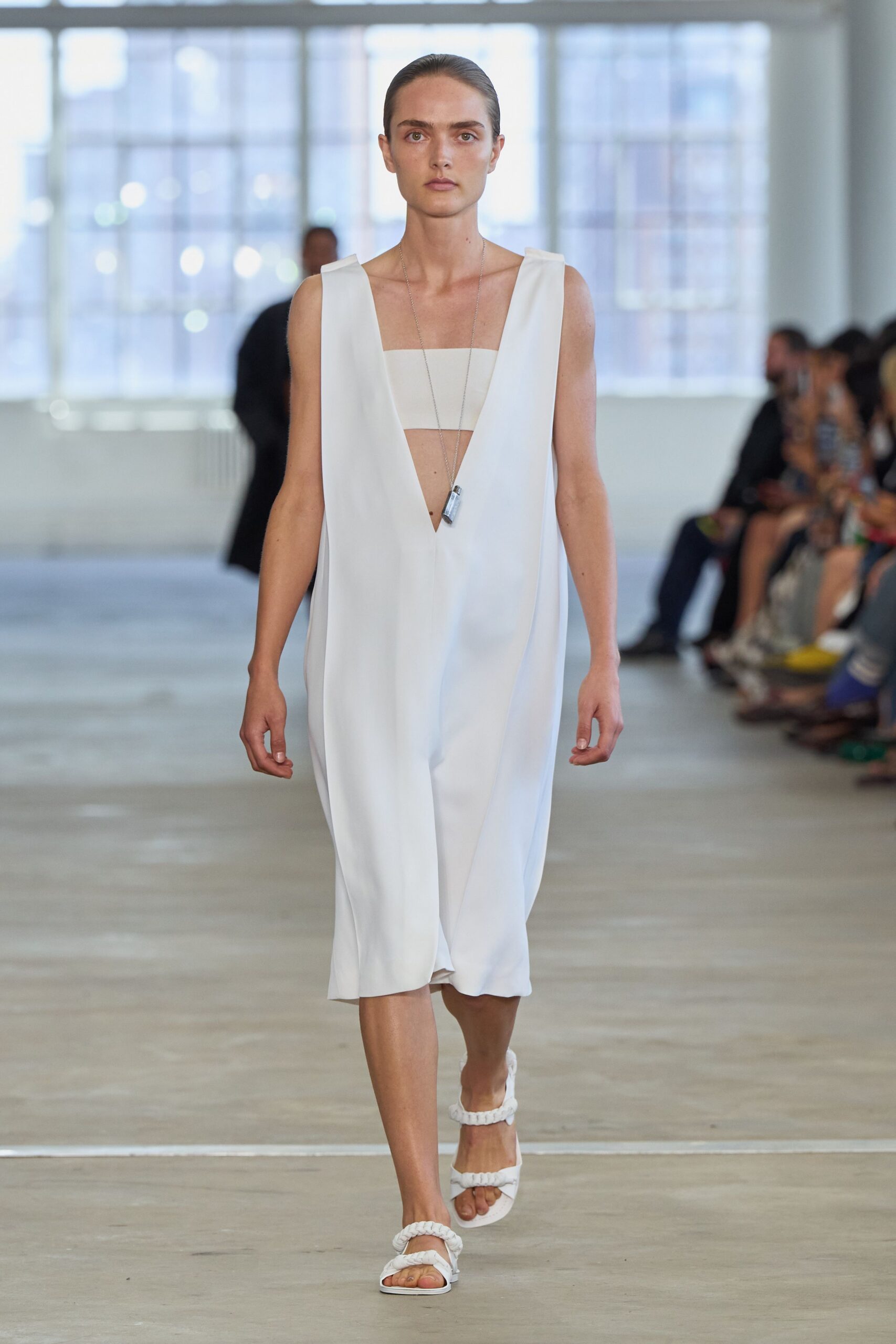 White crepe dress with a plunging V and soft cocoon drape. Straight bandeau panel visible beneath the low opening. Long silver chain with a small vial-shaped pendant. White sandals with thick braided straps across toe and ankle. Crossing a sunlit loft after swimming, concrete still cool underfoot.