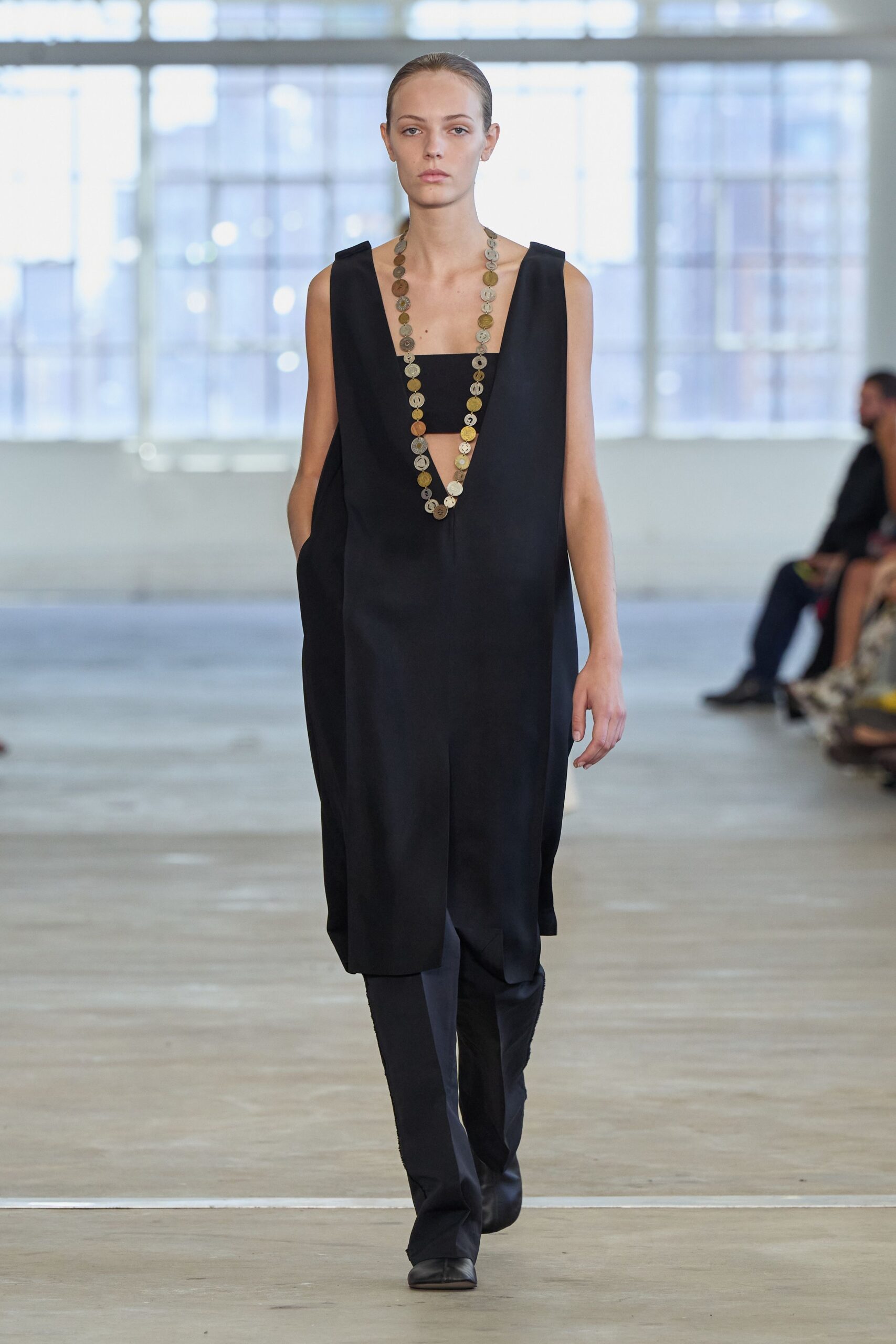 Long necklace of mismatched coin discs, hanging nearly to the waist. Black column tunic with a square bib inset and deep side openings. Black narrow trousers with a satin side stripe and slight break. Pointed black leather boots peeking from under the hem. Museum atrium at opening hour, footsteps carrying on the concrete.