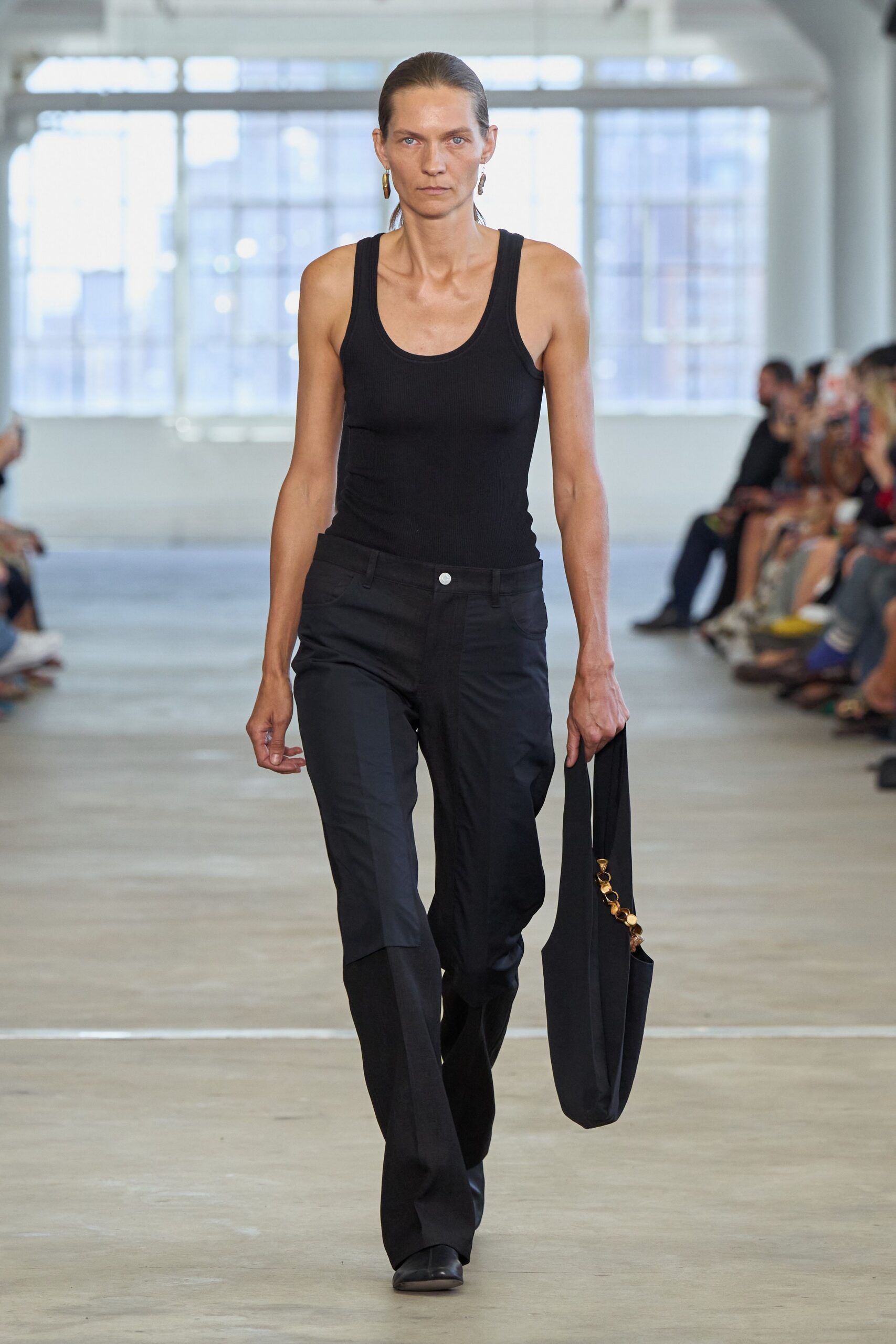Black ribbed tank, cut close with a deep scoop neck. Black low-rise trousers with pressed flares and a pale single button. Soft black tote hung long, strap threaded with amber discs. Mismatched drop earrings in dull gold. Walking into the office on Saturday to water somebody else's plants.