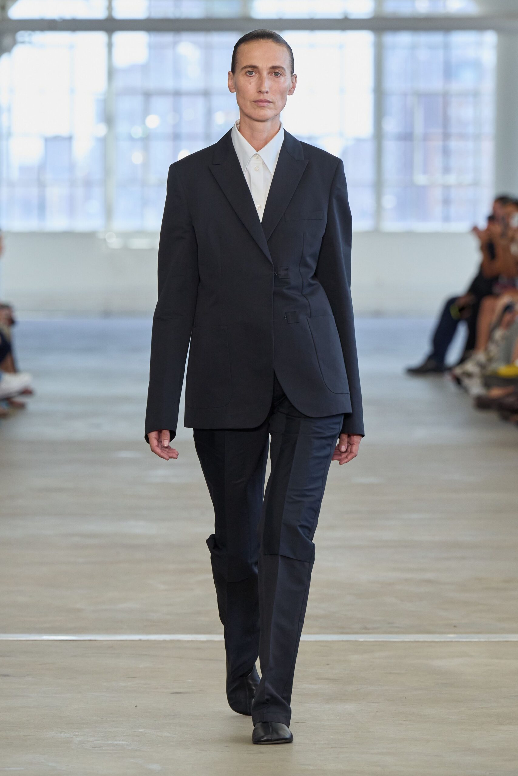 Black suiting blazer cut askew at the waist with low flap pockets. White cotton shirt buttoned clean to the collar. Black tailored trousers with a wide shin panel wrapping one leg. Square-toe black leather boots. Early at the municipal courthouse, carrying papers nobody wants to sign.