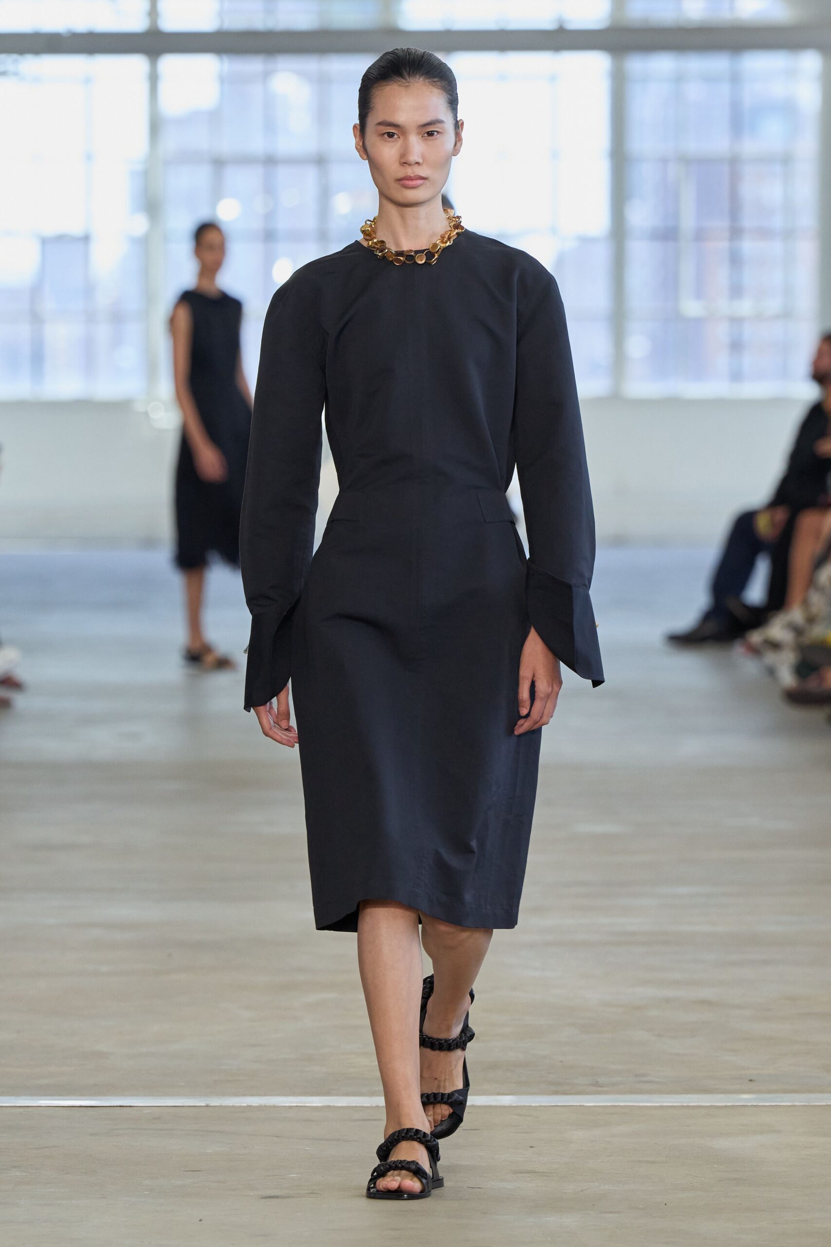 Chunky amber-resin links sitting tight against the collarbone. Black midi dress in crisp matte cloth, dropped waist and split cuffs. Black flat sandals with braided straps crossing the toes. Walking into a museum lecture after rain, marble floors still damp at the doors.
