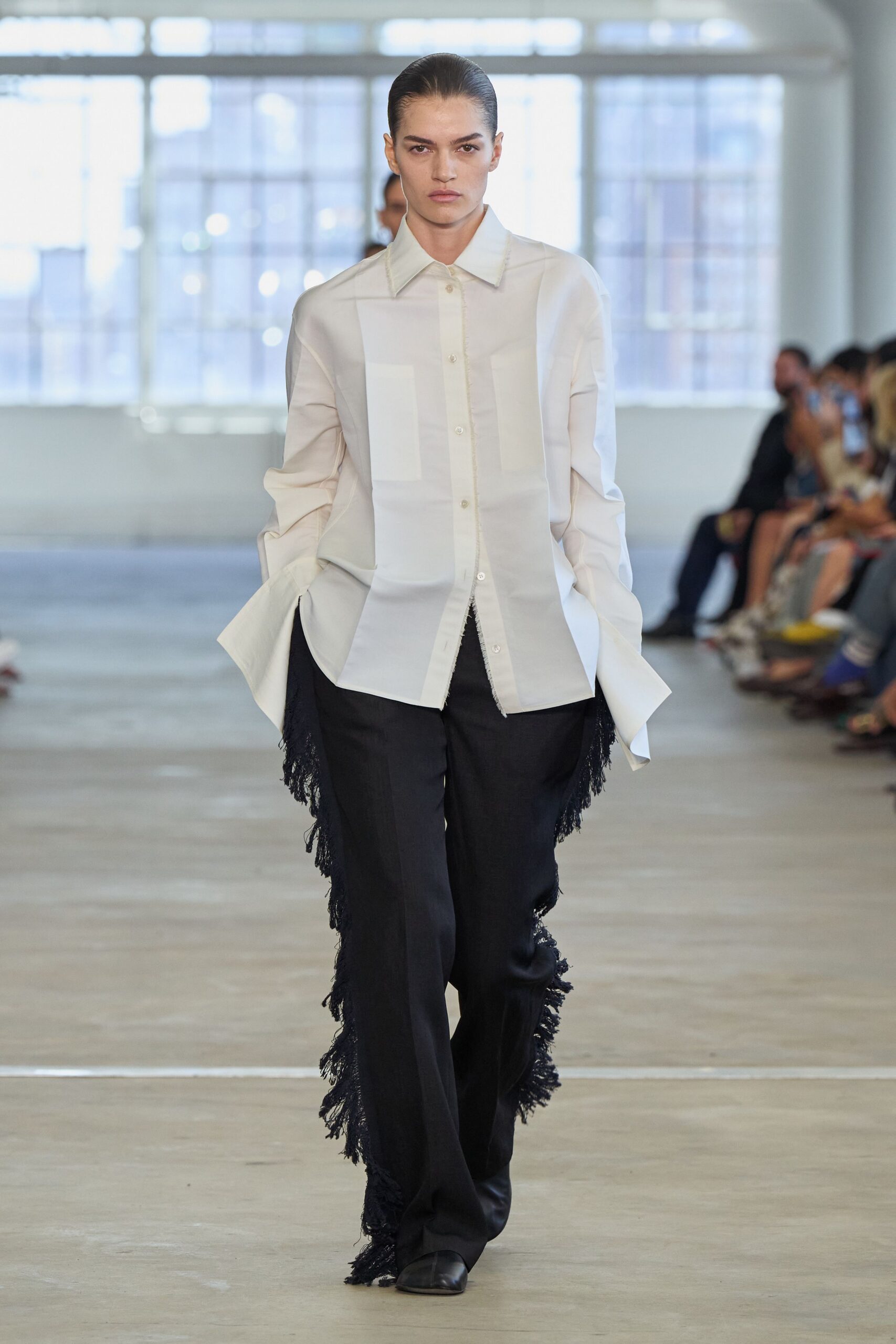 White cotton shirt with dropped shoulders and raw-edged placket seam. Black wide-leg trousers, shaggy fringe running from hip to hem. Square-toe black leather boots. Walking into a sunlit loft meeting before anyone has opened the windows.