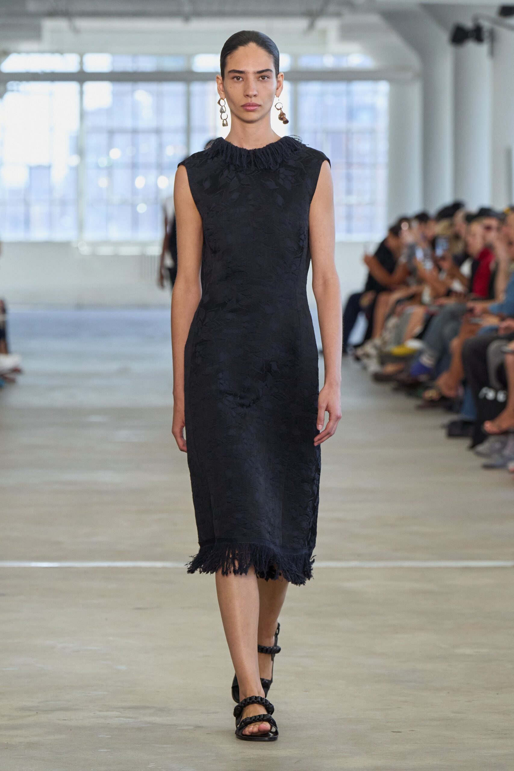 Mismatched drop earrings with linked silver loops and a brown bead. Black floral jacquard column dress, fringed at the neck and uneven hem. Black sandals with thick braided straps crossing the toes and ankle. Walking into a memorial service held in an airy converted mill.