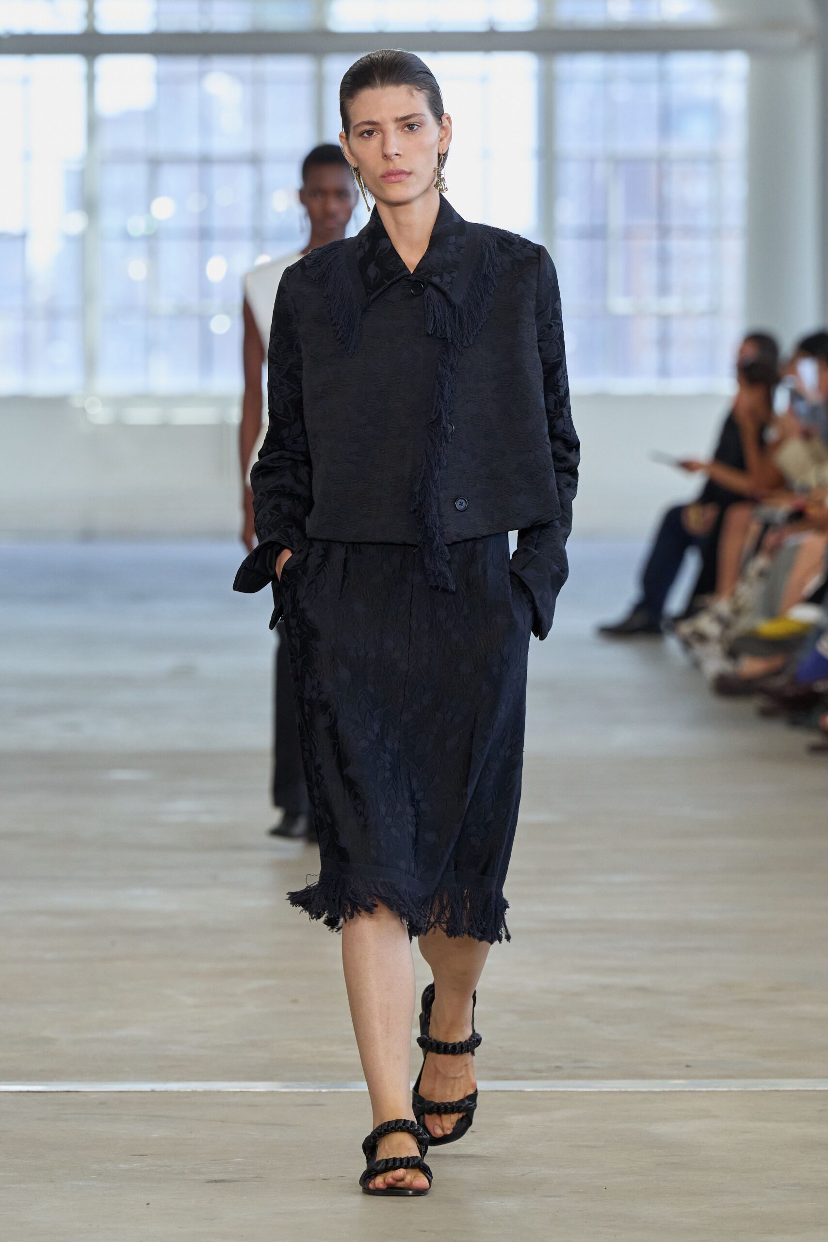 Black floral jacquard jacket with long fringe tracing the collar and placket. Matching midi skirt, fringed hem shaking loose around the knees. Black sandals with padded braided straps. Crossing a museum atrium after rain, still carrying the damp air in.