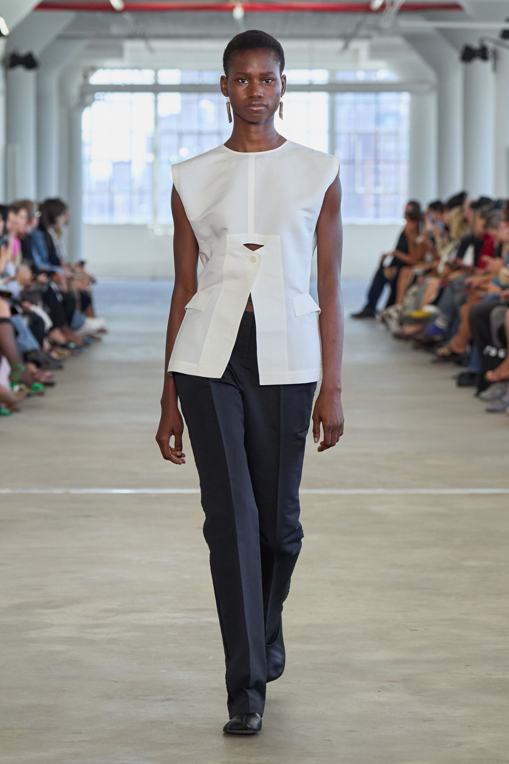 White structured shell top with a skewed placket and triangular cutout. Slim black trousers with a pressed crease and slight boot flare. Black leather square-toe boots. Long bar earrings in brushed metal. For the hour before a panel talk, still carrying your coffee.