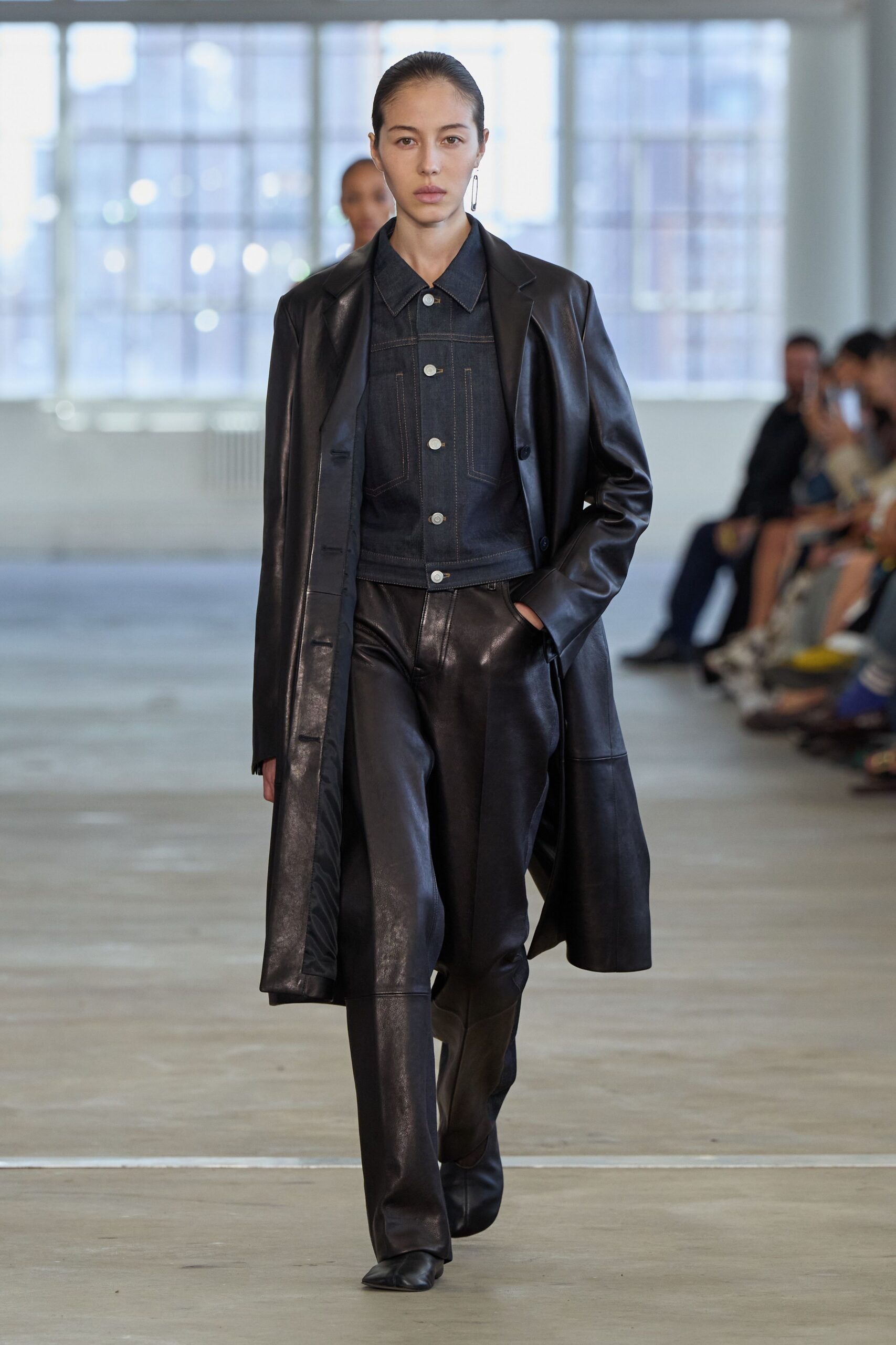 Single elongated silver drop earring. Long black leather coat with a dry, lightly reflective finish. Dark indigo denim jacket, contrast topstitching and pale metal buttons. Black leather trousers falling straight over square-toe boots. Black square-toe leather boots. Crossing an office atrium before anyone else has bad news.