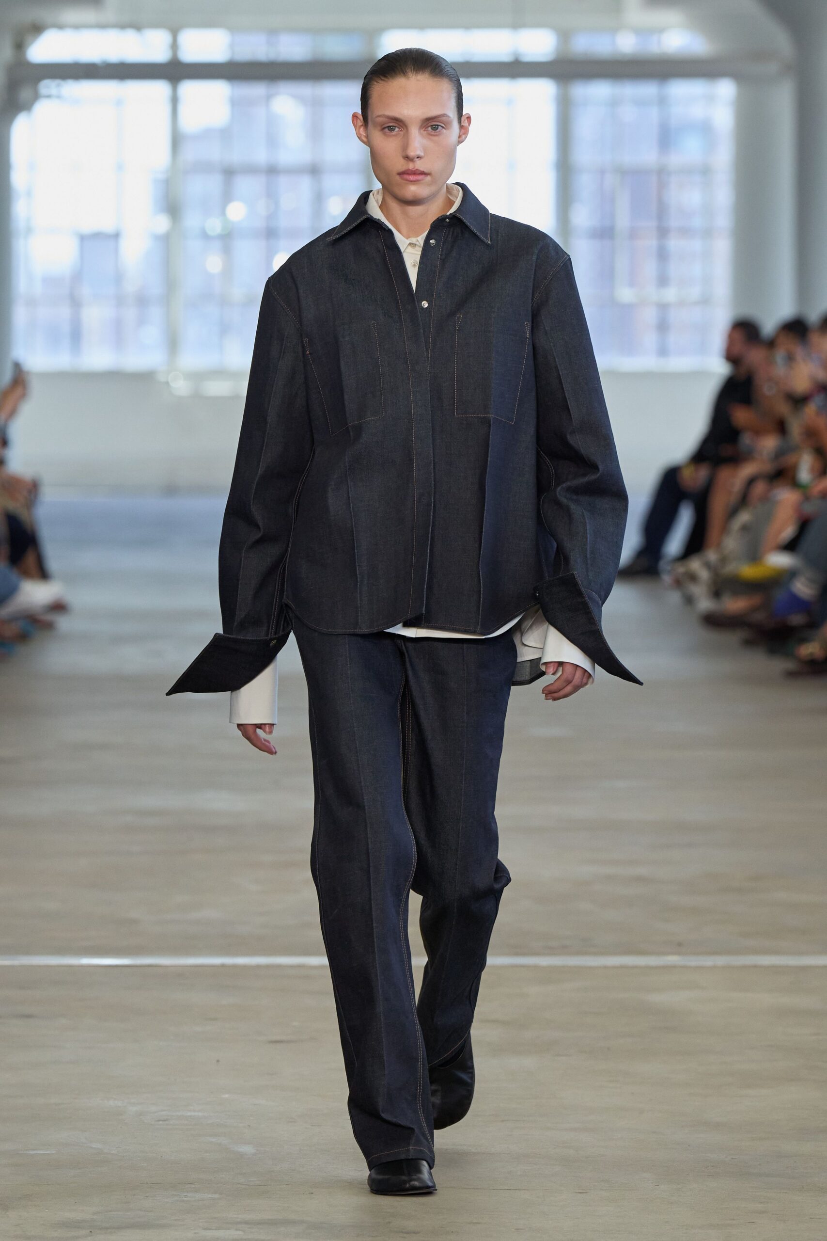 White cotton shirt with long cuffs jutting past the sleeves. Dark raw-denim overshirt, contrast topstitching tracing big square chest pockets. Matching straight-leg denim trousers with a sharp stitched crease. Black leather square-toe boots. First train out after a night spent in someone else's studio.