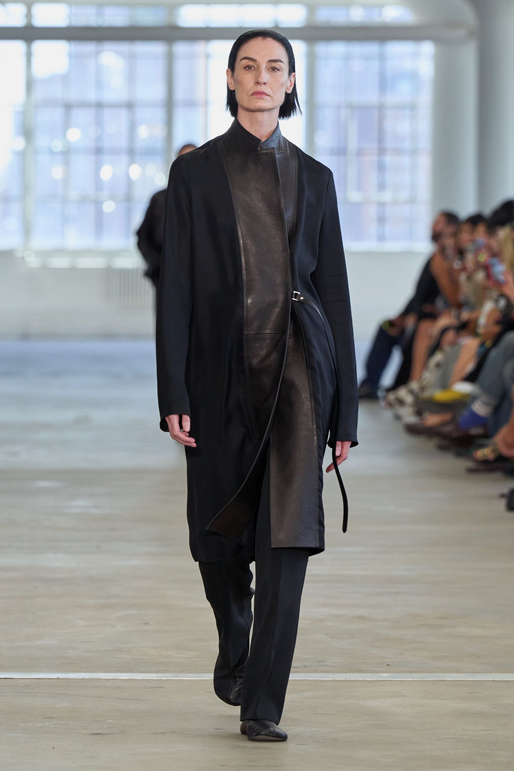 Long black coat with a matte leather front panel and one loose strap. Black straight trousers breaking softly over the shoe. Pointed black leather boots, vamp cut clean and close. For the quiet walk back through office towers after an ethics hearing.
