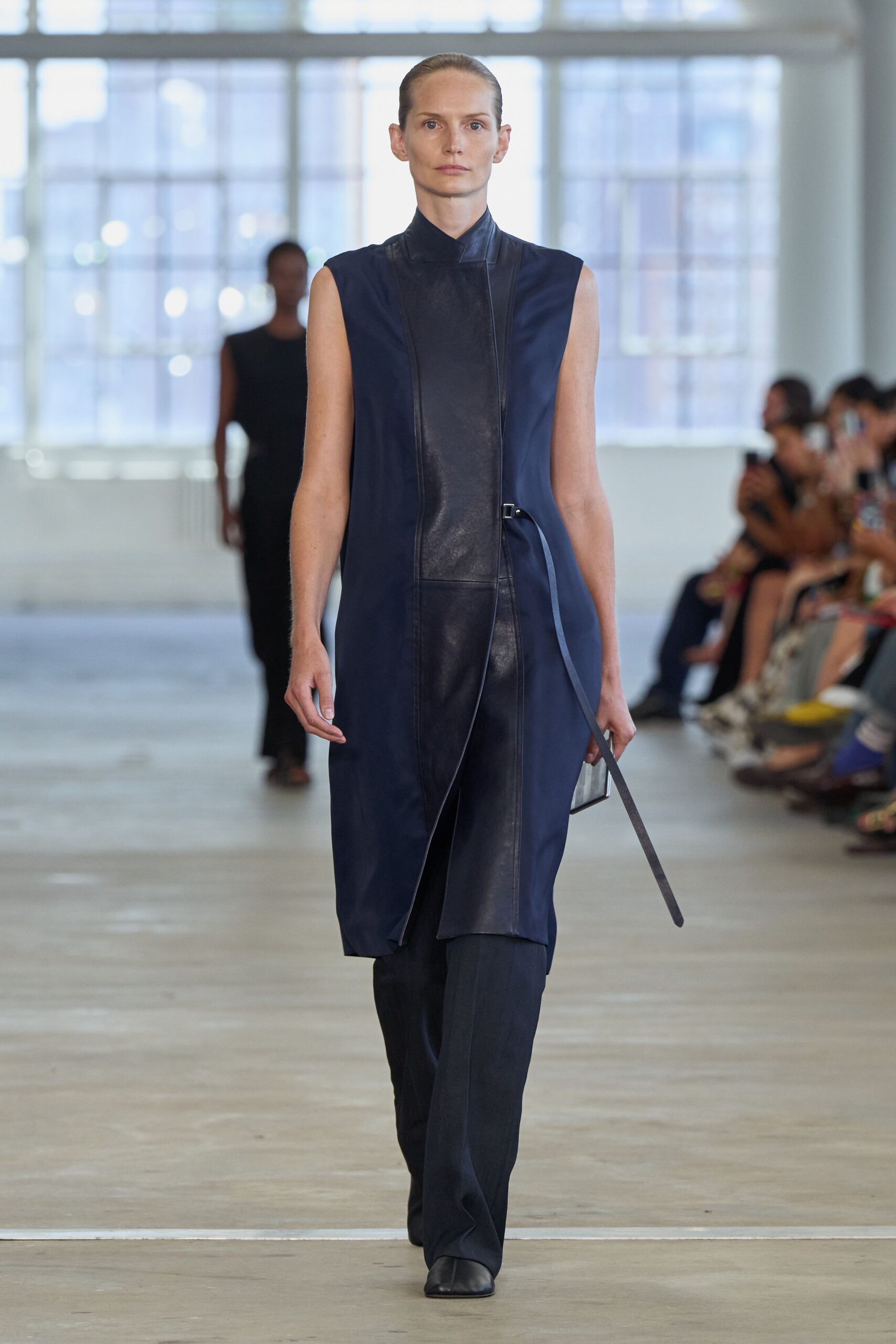 Ink navy sleeveless tunic with a black leather bib and asymmetrical stand collar. Narrow leather strap buckled at the waist, one end hanging past the knee. Dark tailored trousers with a soft break over the shoe. Black square-toe boots. For crossing a glass office atrium before anyone else has arrived.