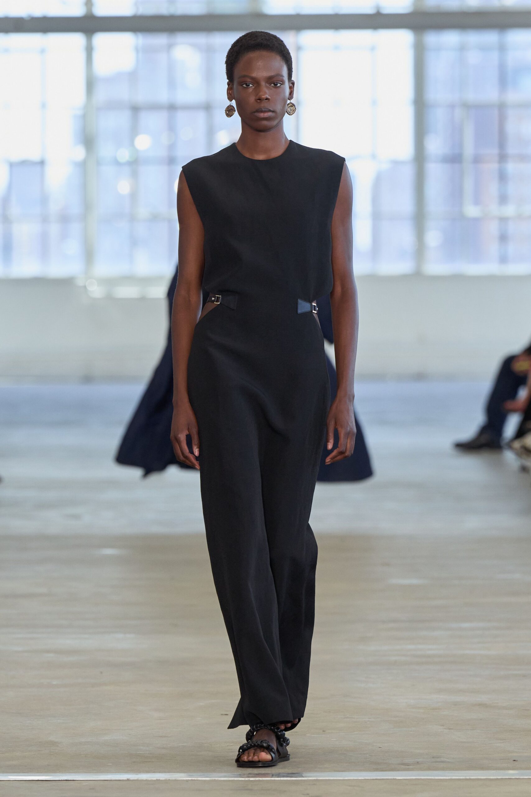 Hammered round drop earrings in dull gold metal. Black sleeveless jumpsuit with waist cutouts cinched by tiny side buckles. Flat black sandals with knotted straps across the toes. Walking across a quiet museum atrium before the doors unlock.
