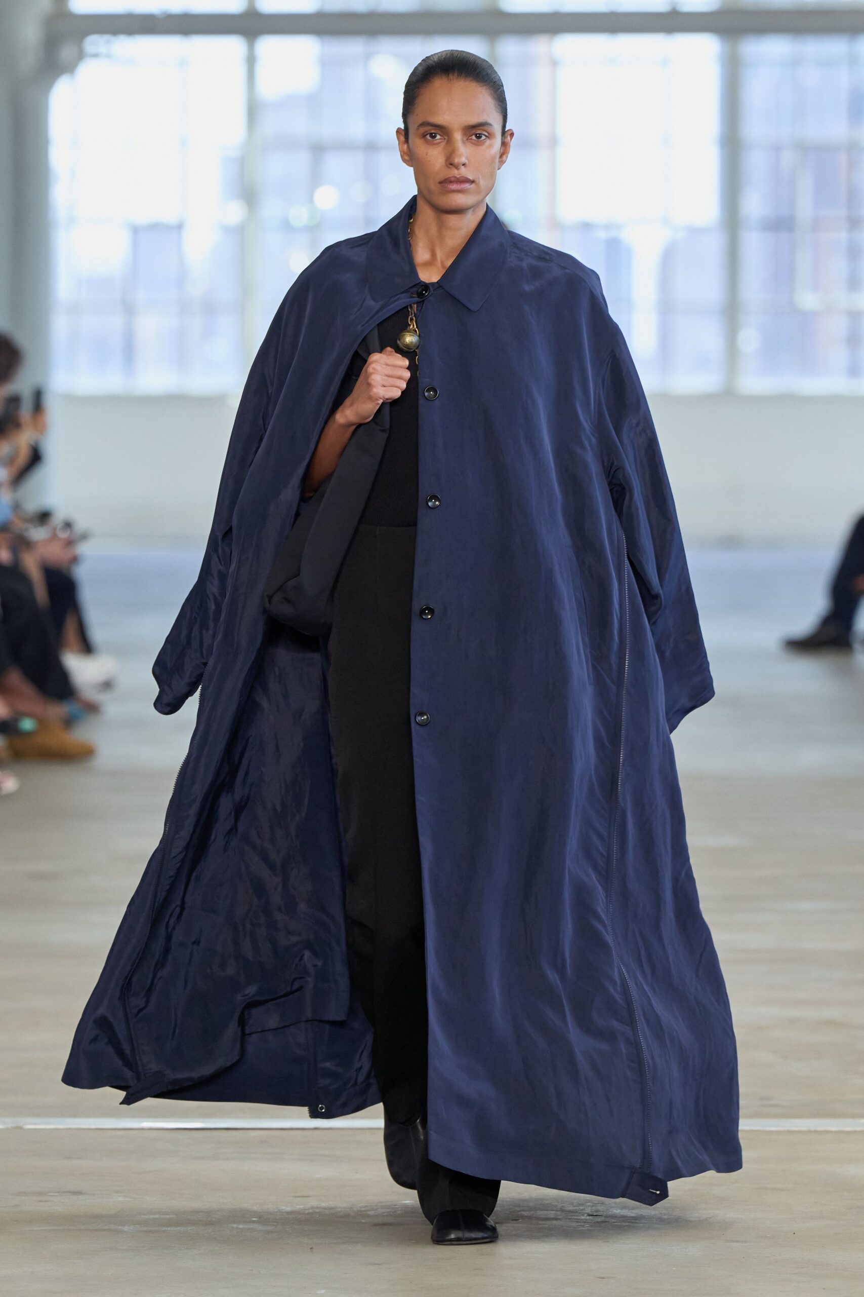 Ink-blue coat in crisp technical cloth, buttoned high with zip-open side seams. Black knit top, cut close at the throat. Black wide trousers falling straight under the coat. Square-toe black shoes. Brass orb pendant caught at the placket. Crossing a windy plaza with your keys still in your fist.