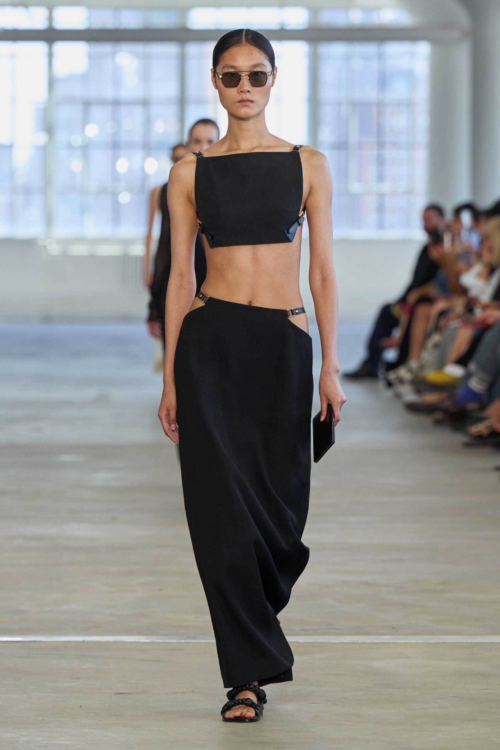 Narrow brown-tinted rectangular lenses with slim gold arms. Black cropped bandeau, clipped at the shoulders and side seams. Low-slung black column skirt, hip cutouts held by tiny buckles. Black braided leather sandals. Flat black clutch in hand. Crossing a hotel lobby before anyone has set down their iced coffee.