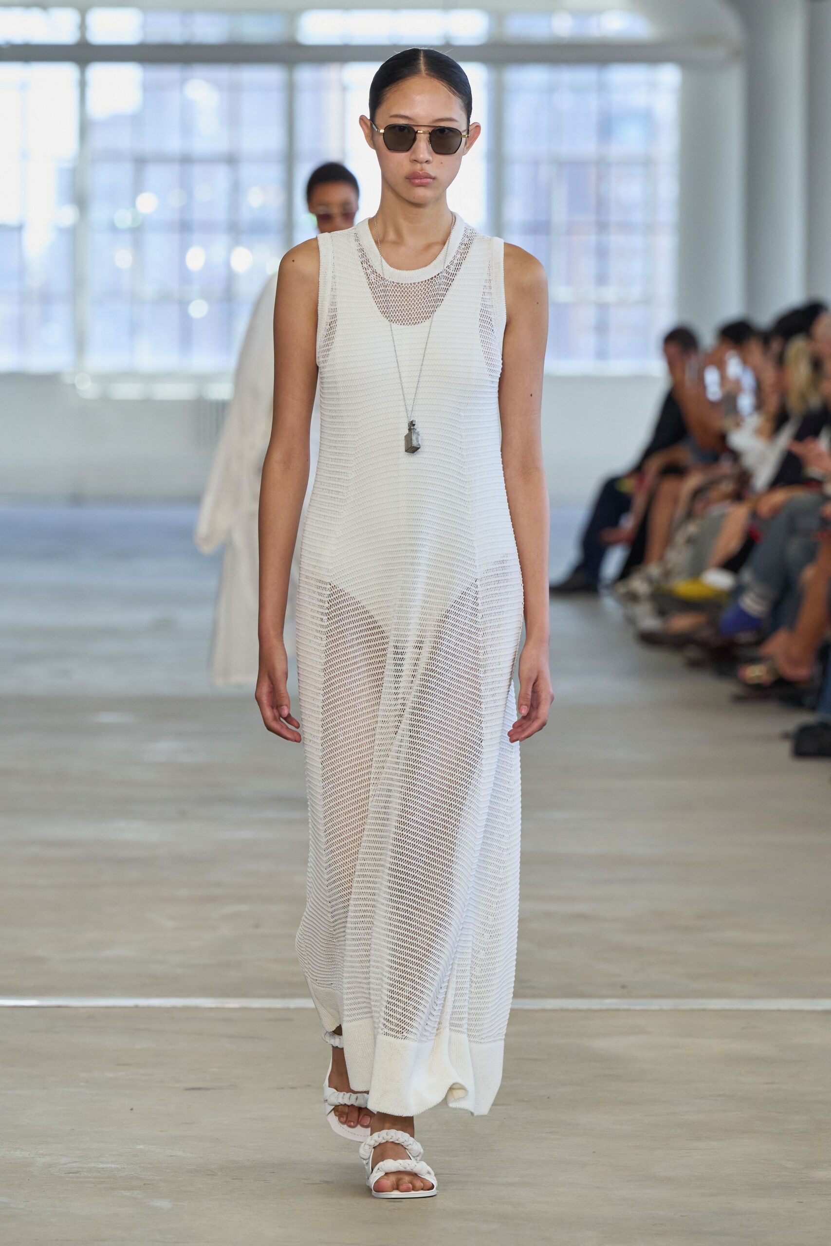 Square smoke lenses in a thin silver wire frame. Long chain with a rough grey stone pendant. White open-knit tank dress, denser ribbing at the hem. White wide trousers ghosting through the mesh. White padded thongs with ruched straps. Walking the hotel corridor before breakfast, still damp from the pool.