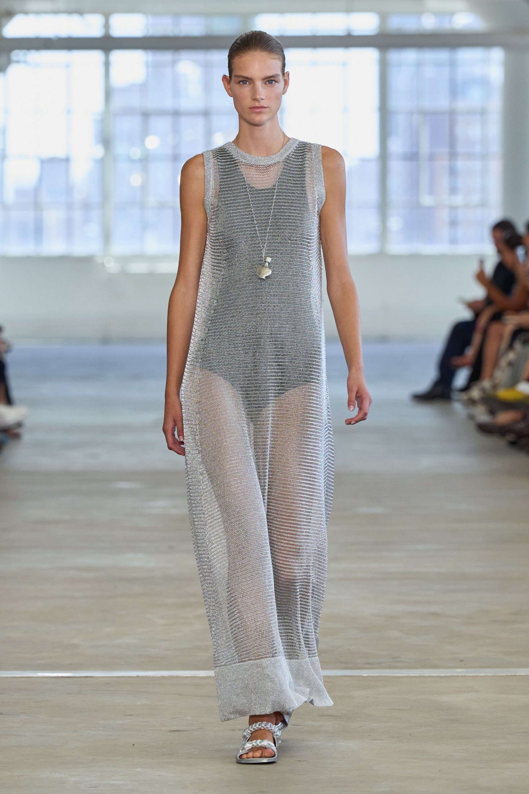 Floor-length silver mesh dress, open laddered knit over a dark slip. Long cord necklace with a small brushed metal pendant. Silver flat sandals with puffed braided straps. Walking across a museum atrium while the fountains are switched off.
