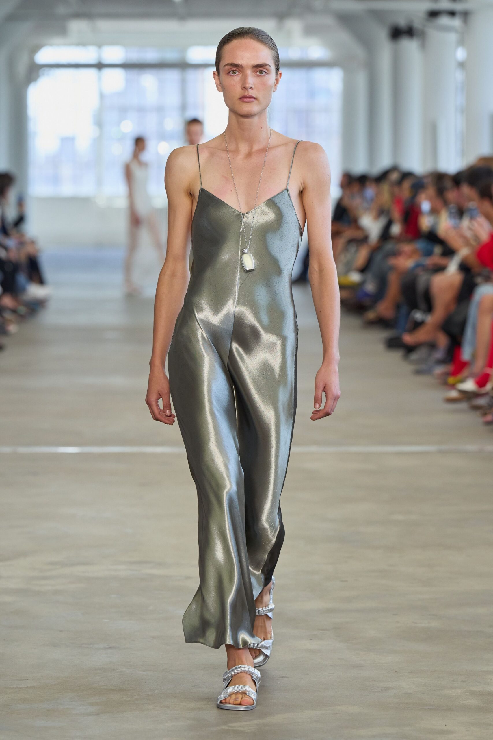Liquid pewter satin jumpsuit with pin-thin straps and a sharp center seam. Long silver chain with a small milky rectangular pendant. Silver sandals with braided straps across the toes and instep. Crossing a hotel atrium before breakfast service has properly started.