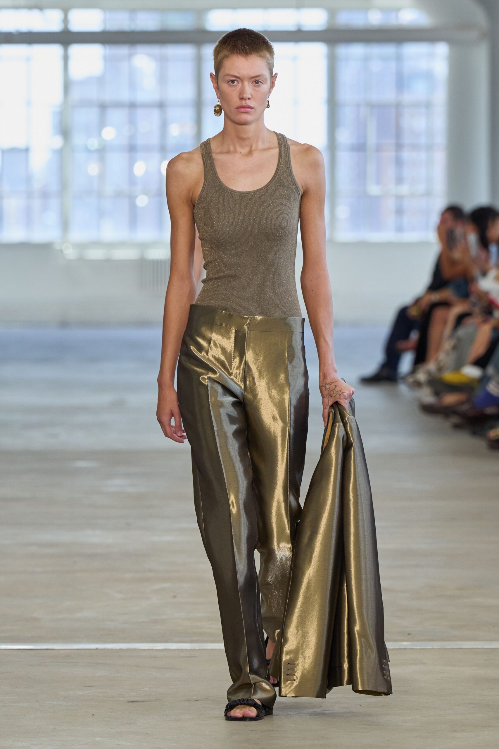 Taupe ribbed tank, cut close with a scooped athletic neck. Bronze-coated trousers with a liquid shine and creased front seam. Matching bronze coat carried loose, vent tabs peeking near the hem. Black braided sandals. Mismatched brass drops. For crossing a near-empty office after midnight, jacket over one fist.
