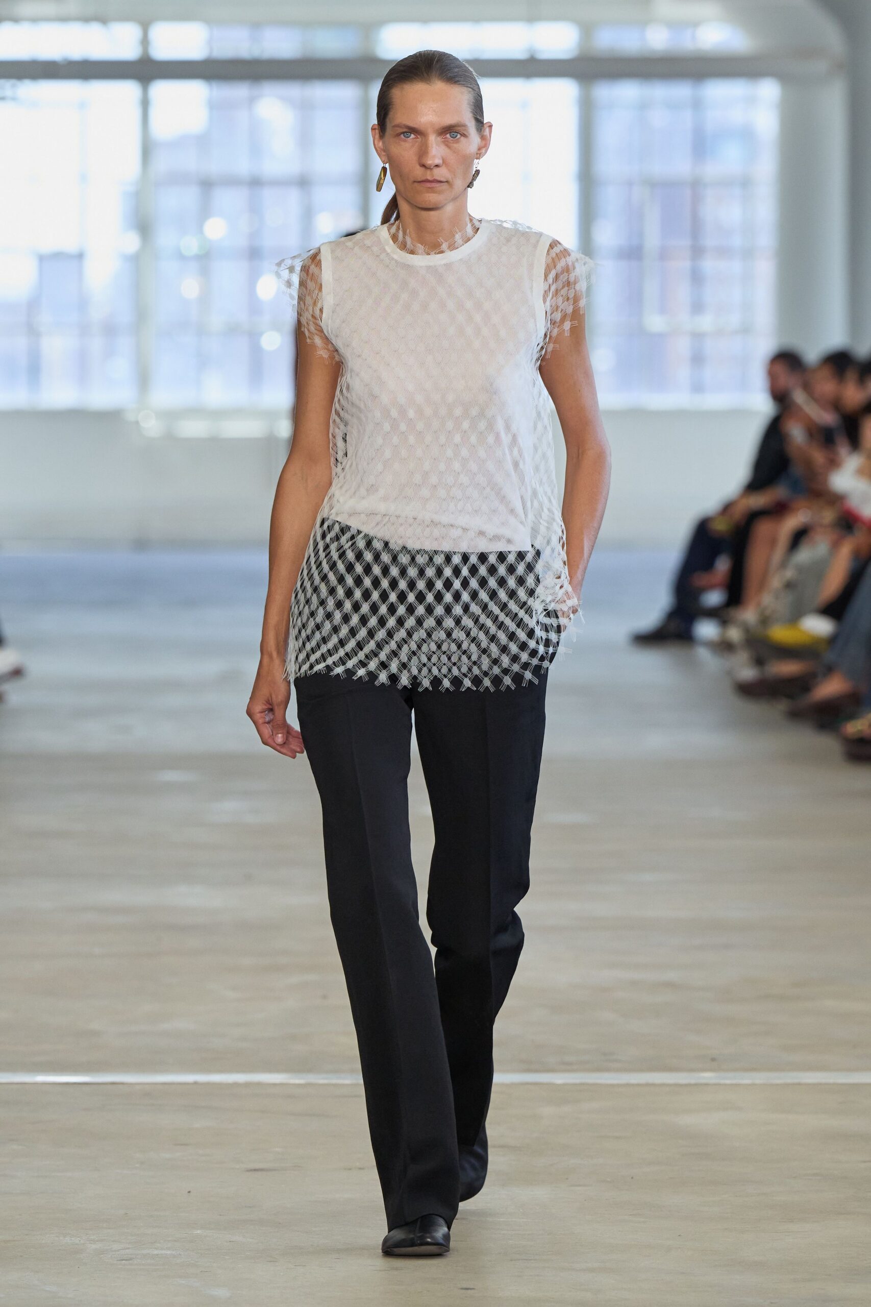 White diamond-mesh shell over a fitted underlayer, fringe needling off sleeves and hem. Black tailored trousers cut straight and long through the shoe. Mismatched brass drops. Walking across an empty civic atrium before the offices fill.