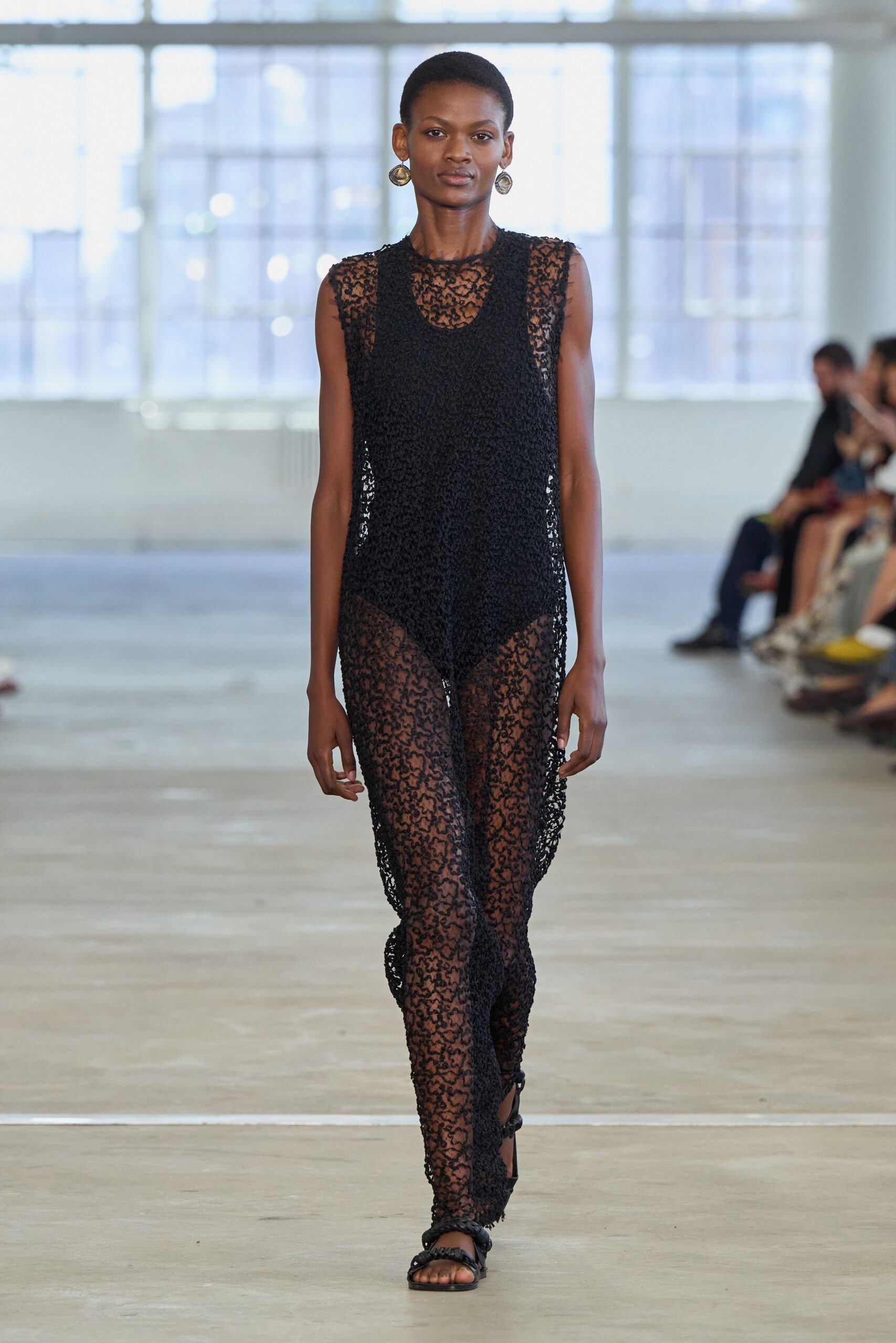 Mismatched silver disc earrings with a hammered, off-center swing. Sheer black looped crochet column, tank-shaped over a denser central panel. Flat black sandals with thick braided straps crossing the toes. Walking through an old swimming hall after the water's been drained.