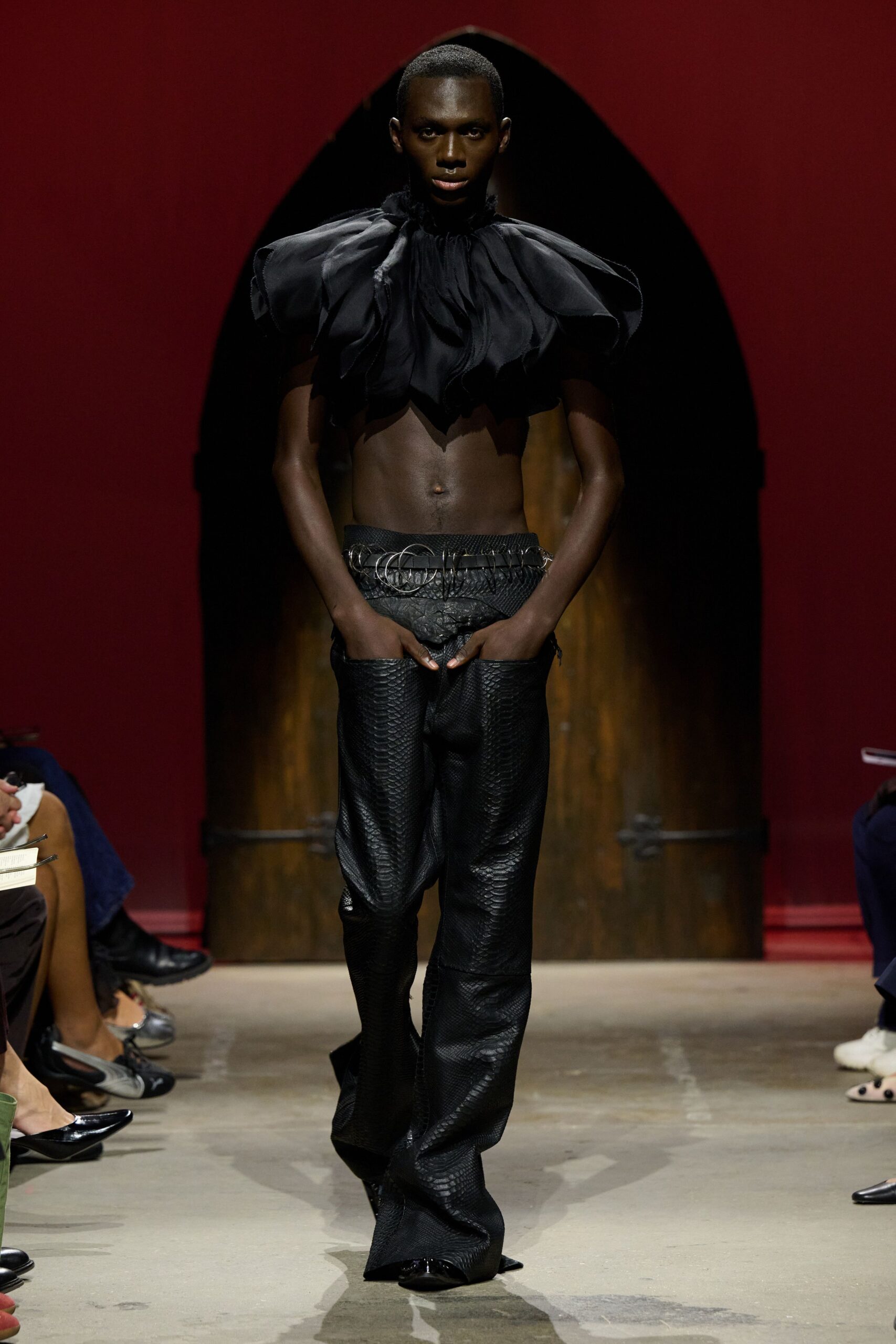 Black chiffon cropped top with swollen petal sleeves and a ruched throat. Black belt threaded with oversized silver rings across the low waist. Black python-embossed trousers slung low, pooling over pointed shoes. For crossing a candlelit dining room after everyone else has sat down.