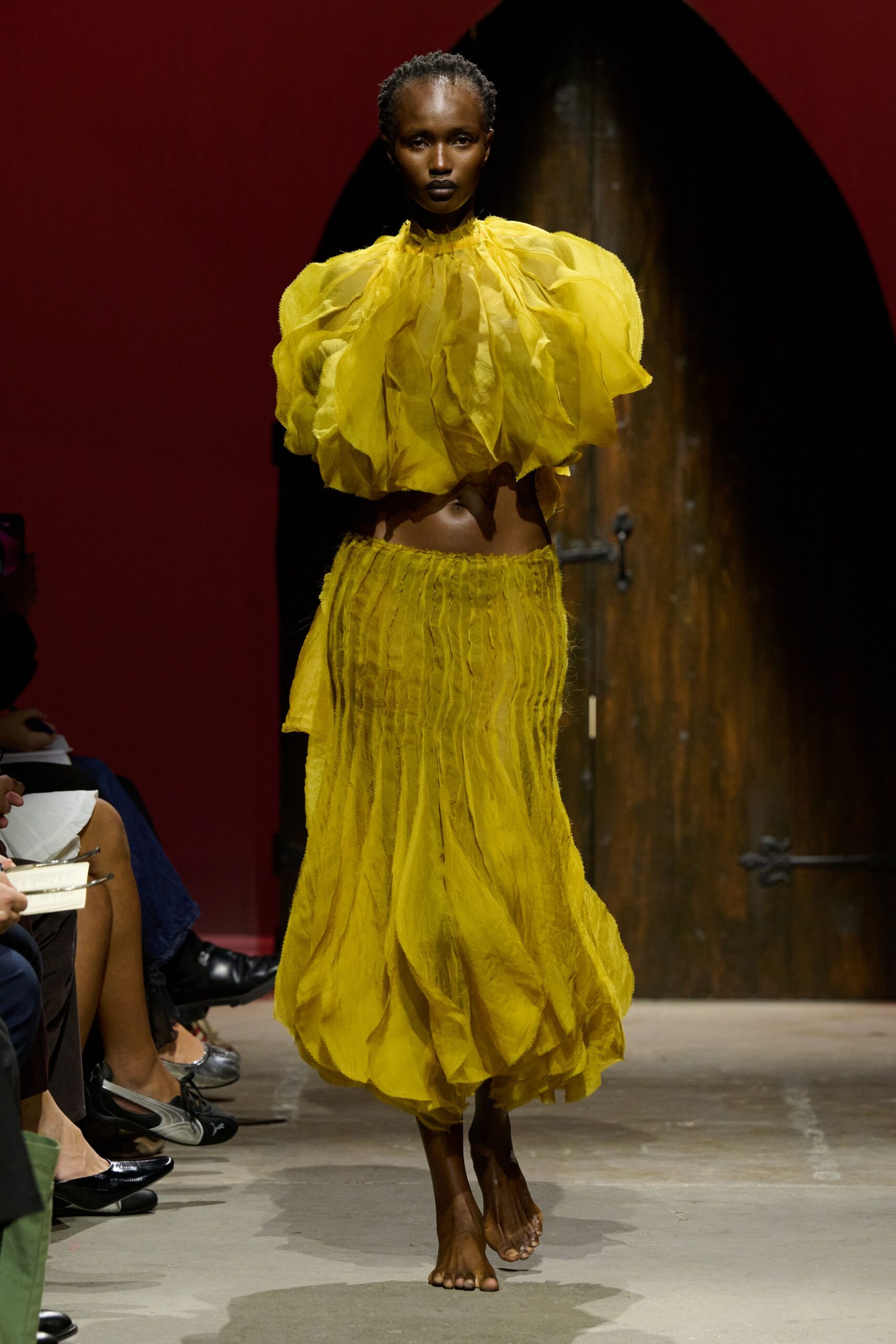 Marigold organza blouse ballooning outward in torn petal-like layers. Matching crinkled skirt, vertically ruched and puffed into a cocoon hem. No shoes, toes fully exposed. Crossing a tiled courtyard just after the fountain was turned off.