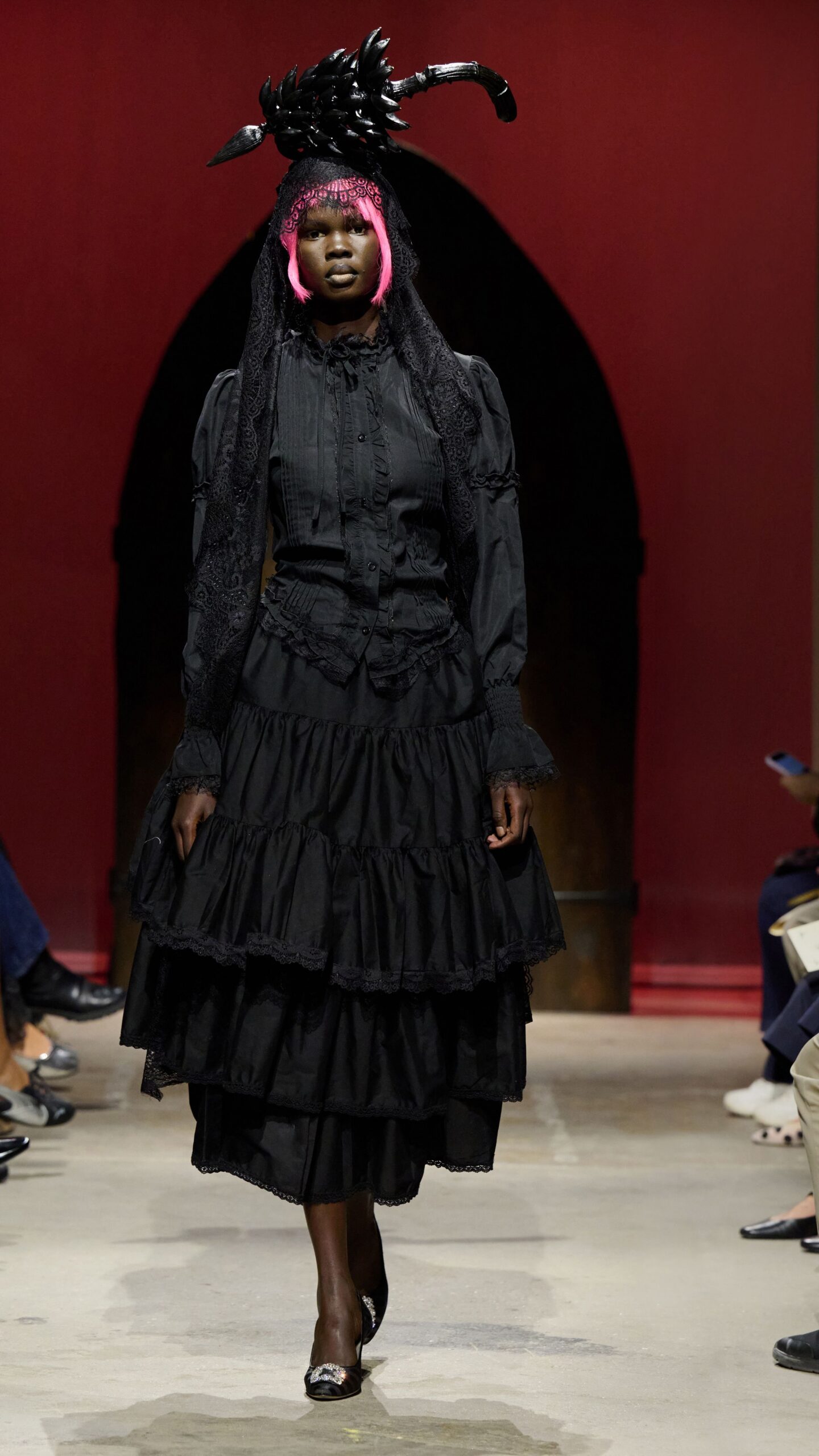 Black sculpted bird headpiece over a long floral-lace veil. Black pintucked blouse with ruffled placket and shirred cuffs. Tiered black cotton skirt, each flounce edged in narrow lace. Black satin mules with a rhinestone buckle at the peep toe. Crossing the churchyard after rain, still wearing the mourning veil.